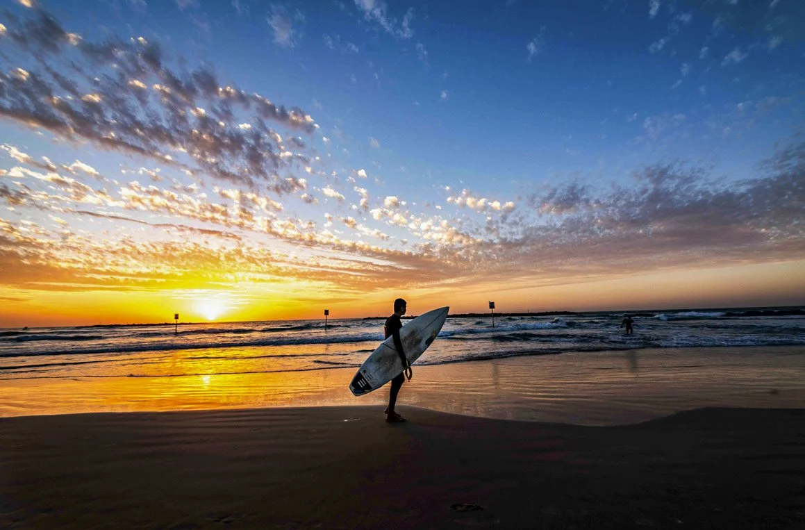A person holding a surfboard on the beach during sunset with a colorful sky and calm ocean waves.