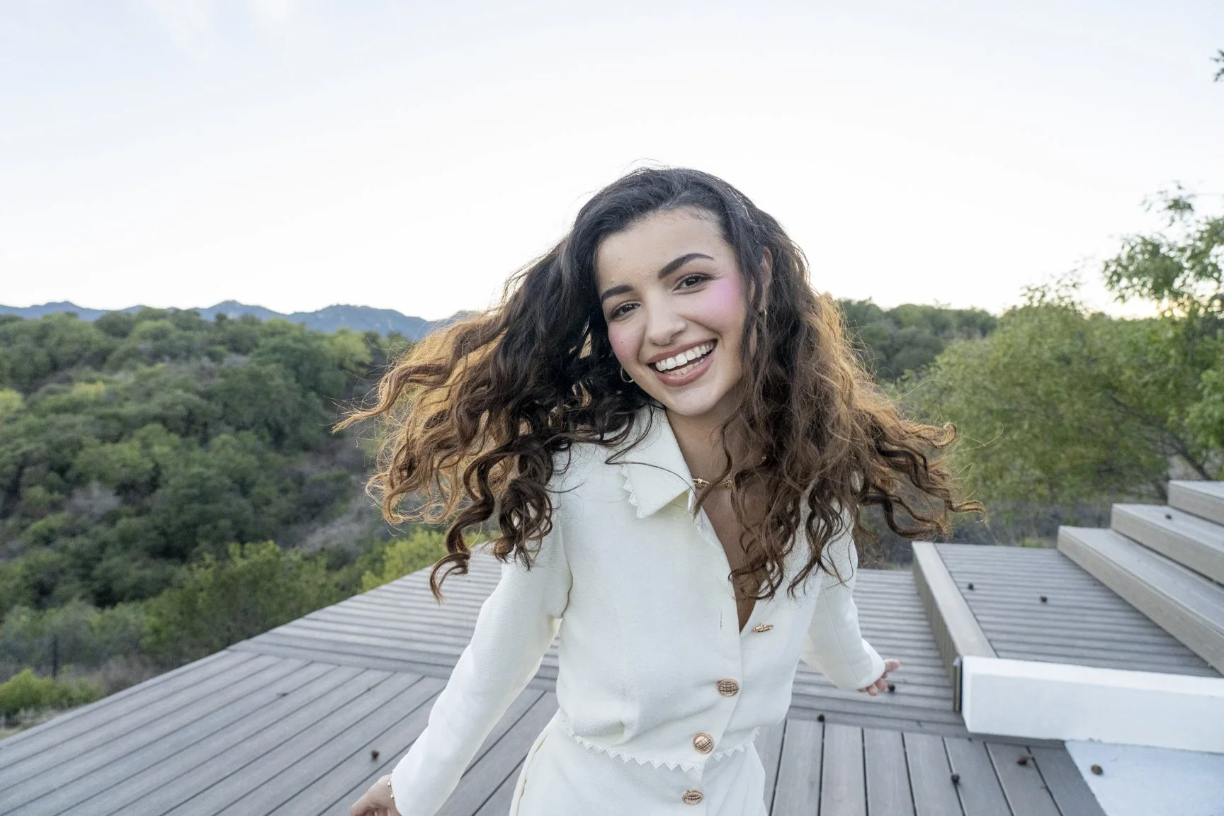 A young woman with long, curly brown hair smiling and leaning on a wooden deck railing outdoors with green trees and distant mountains in the background.