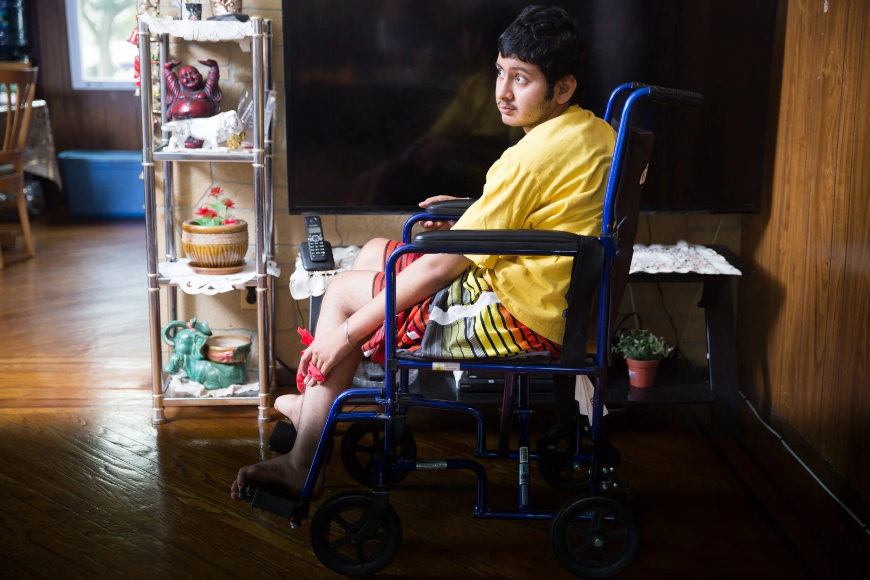 A young man in a wheelchair with a yellow shirt and striped shorts sitting in a living room near a TV, with ornaments on a shelf nearby.