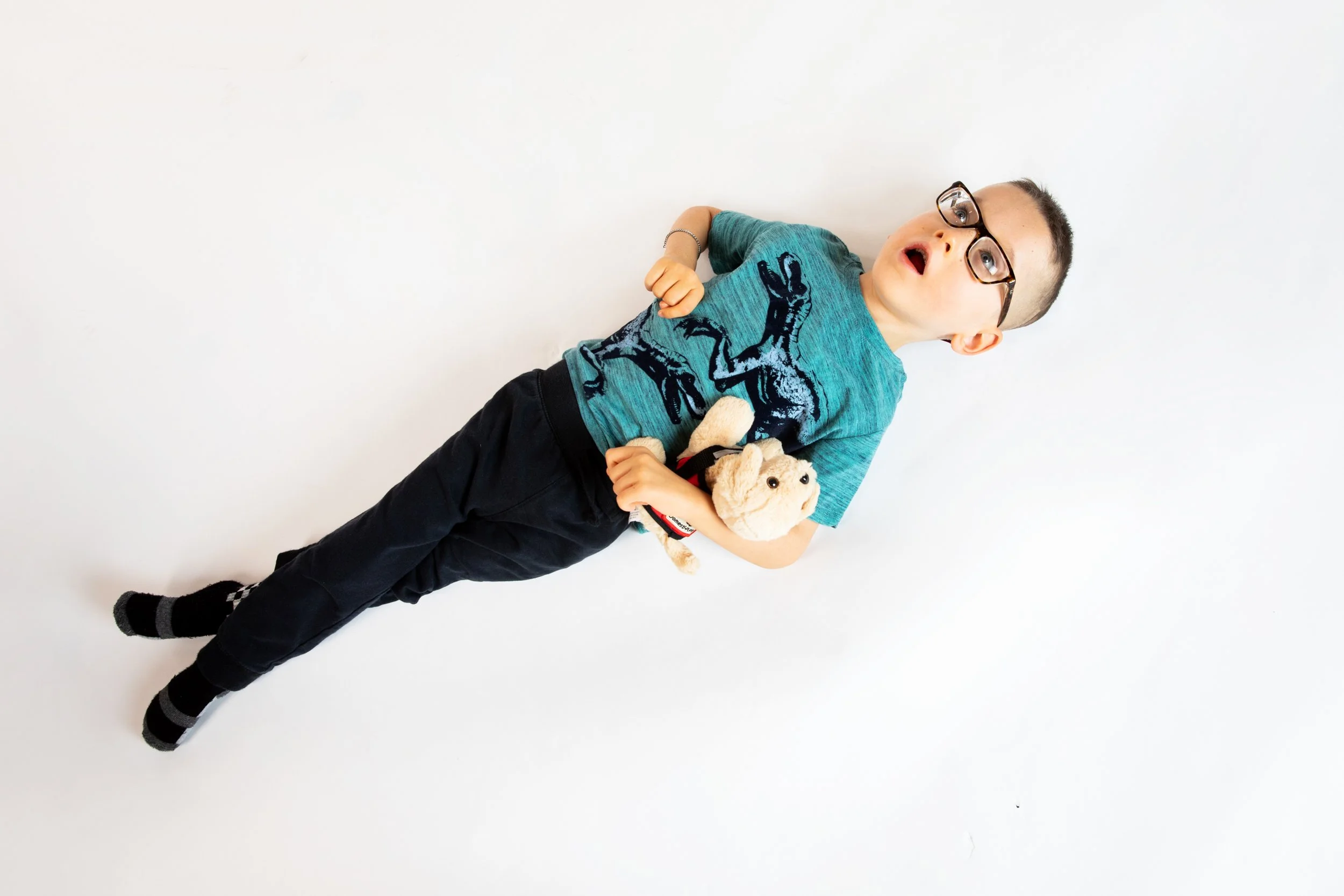 A young boy wearing glasses, a blue t-shirt with dinosaur graphics, and black pants, lying on a white surface, holding a stuffed animal.