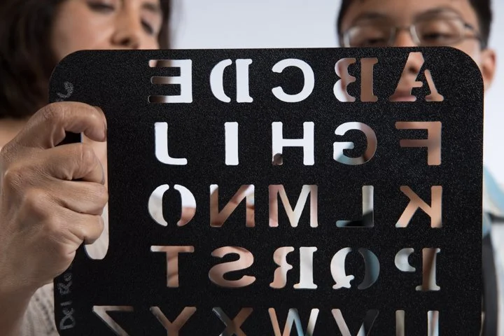 Two people hold up an eye chart with black letters on a white background, partially obscured by a black stencil.