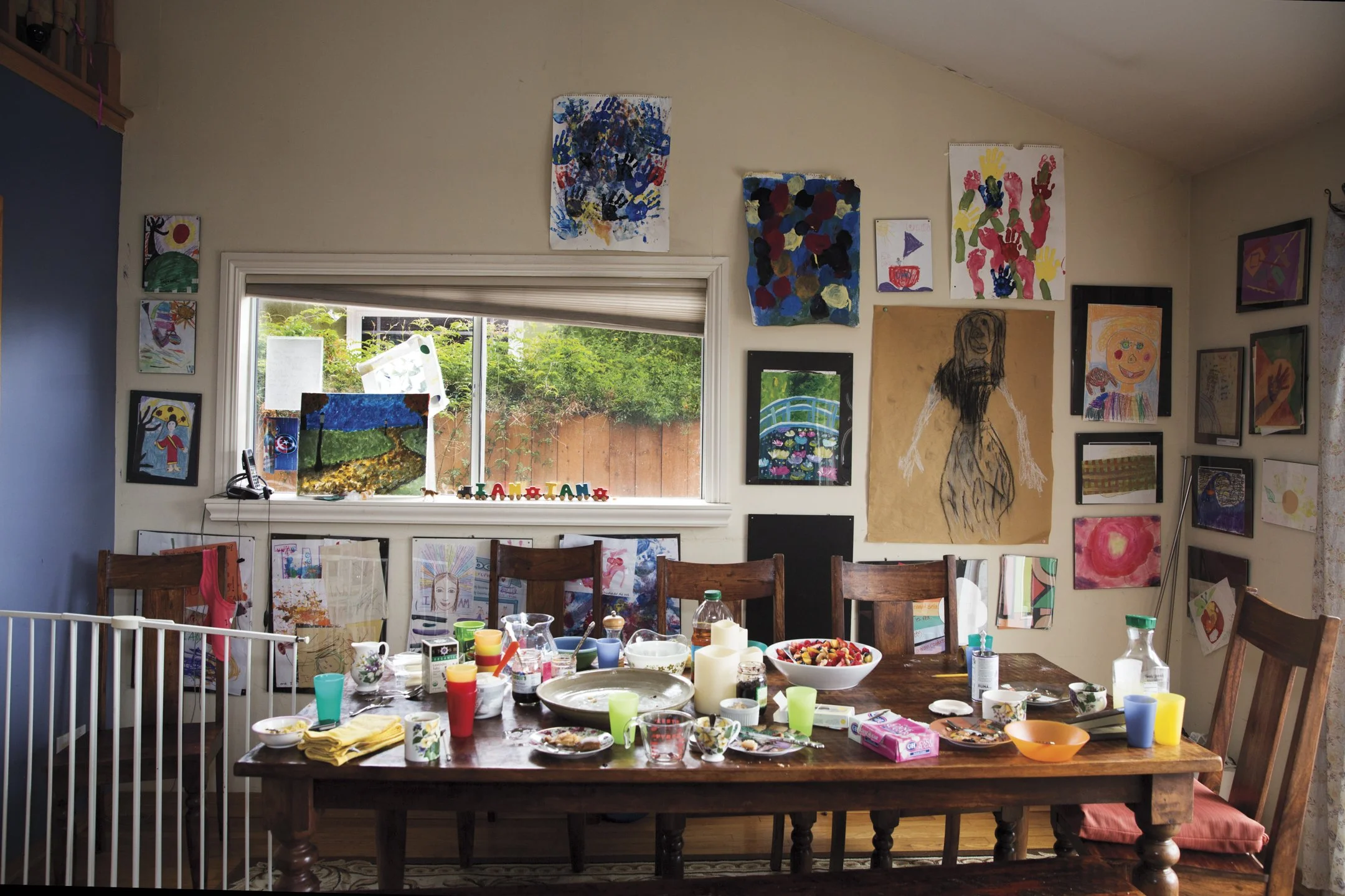 A cluttered dining room with a wooden table filled with dishes, cups, and food remnants. The wall behind is decorated with children's artwork, and a window shows a backyard with a wooden fence and greenery.