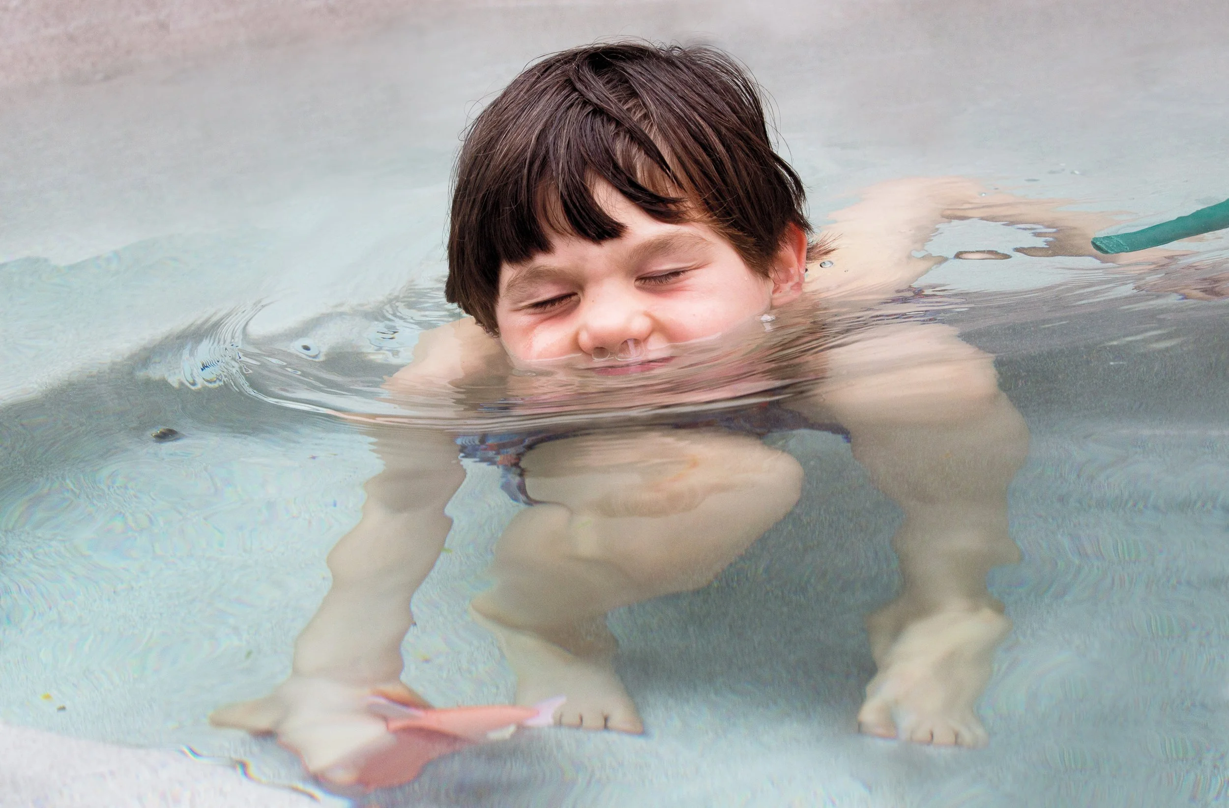 Young boy with brown hair swimming in a pool, resting on his arms with his face partially submerged, eyes closed, enjoying the water.