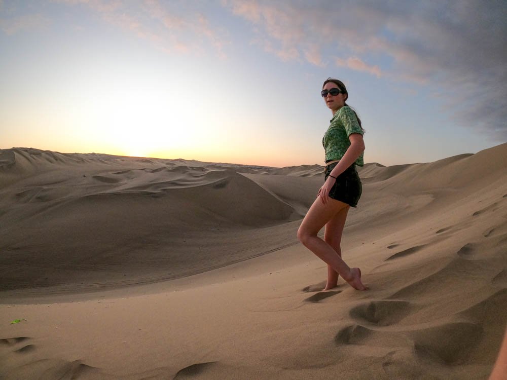 A woman with sunglasses wearing a green top and black shorts standing barefoot in the sand dunes at sunset.