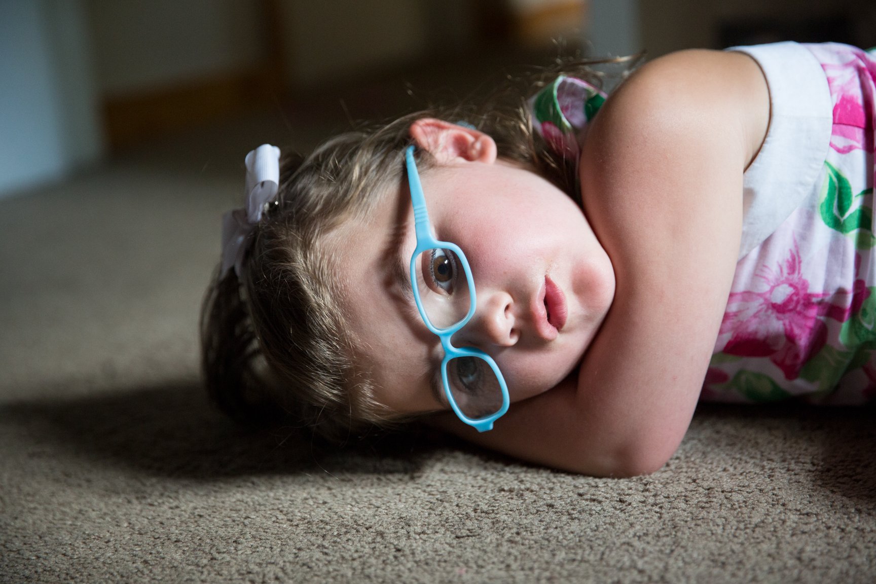 Young girl with glasses lying on her side on a carpeted floor, resting her head on her arm, with a thoughtful or surprised expression.