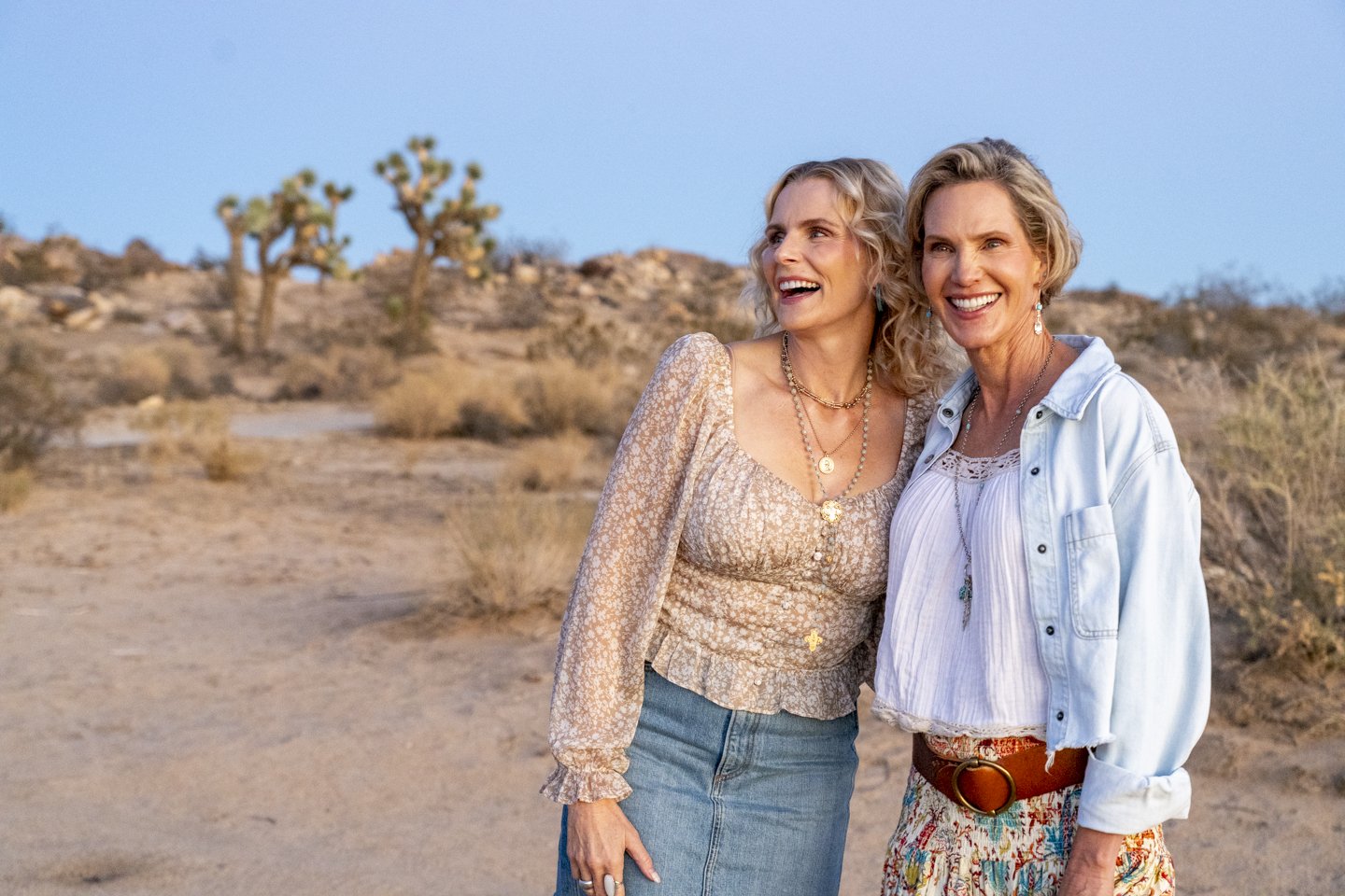 Two women smiling and standing close together in a desert landscape with desert plants and trees in the background, during sunset or late afternoon.