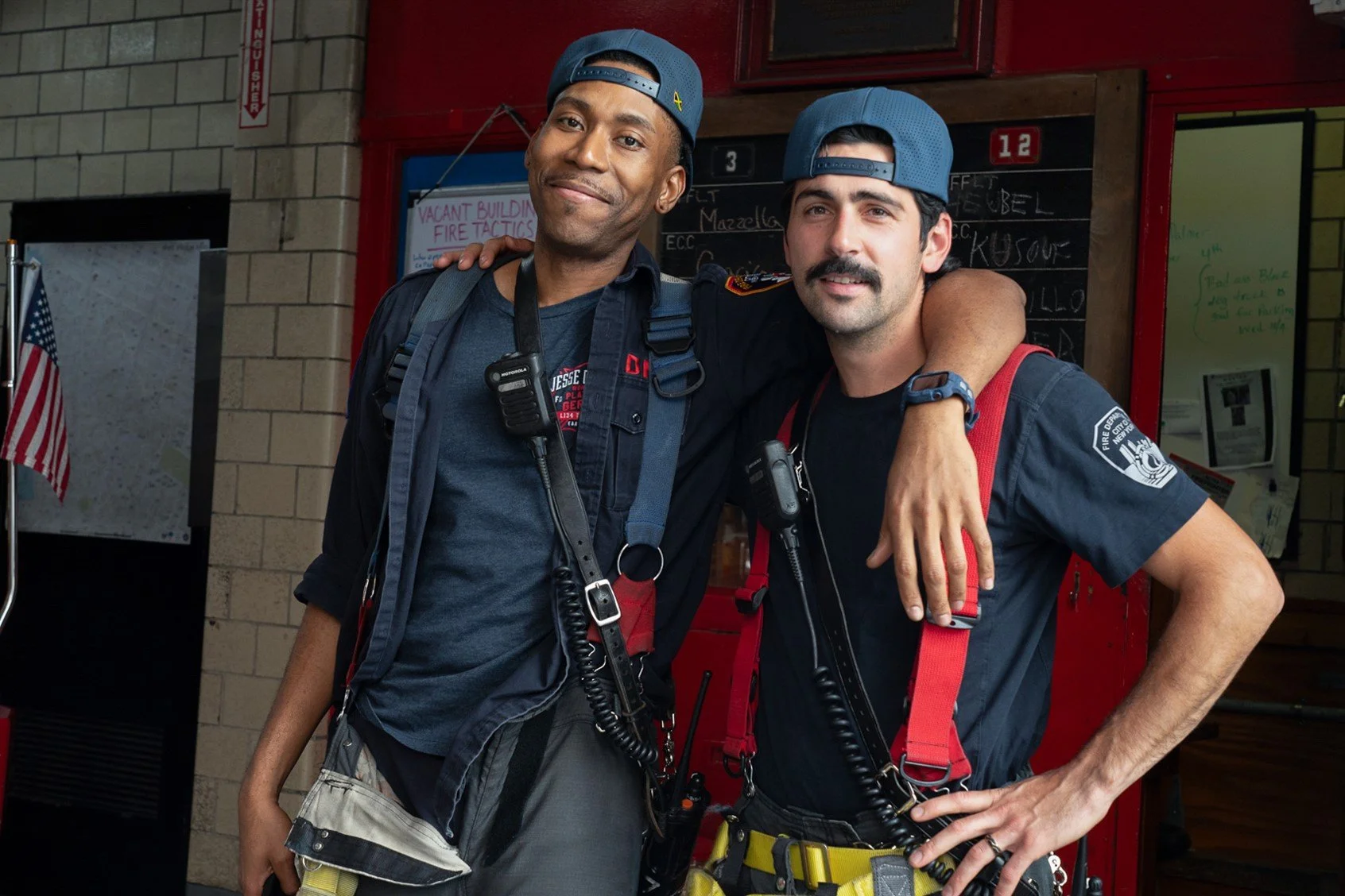Two male firefighters in uniform standing together, smiling, with one arm around each other's shoulders, inside a fire station.
