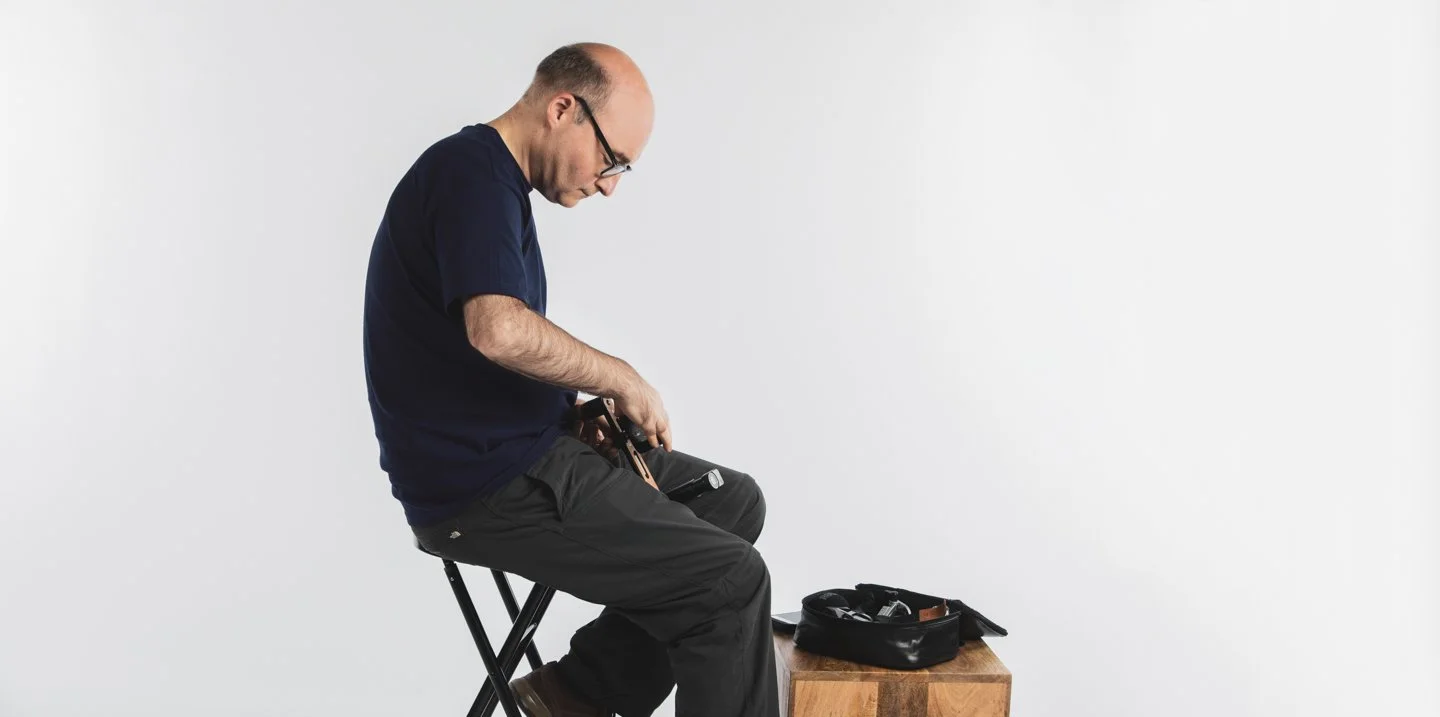 Man sitting on a stool, looking through a small black bag with various tools inside, on a wooden table against a plain white background.
