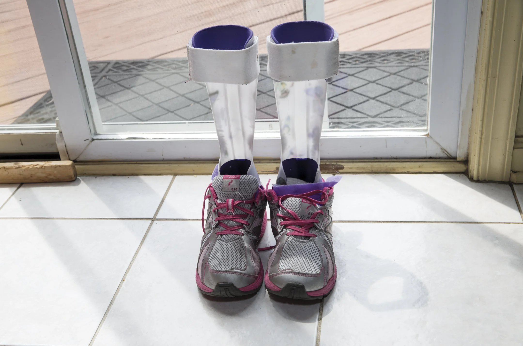 Pair of athletic shoes with pink laces placed indoors on tiled floor, positioned in front of glass door, with rain boots behind them visible through the glass.