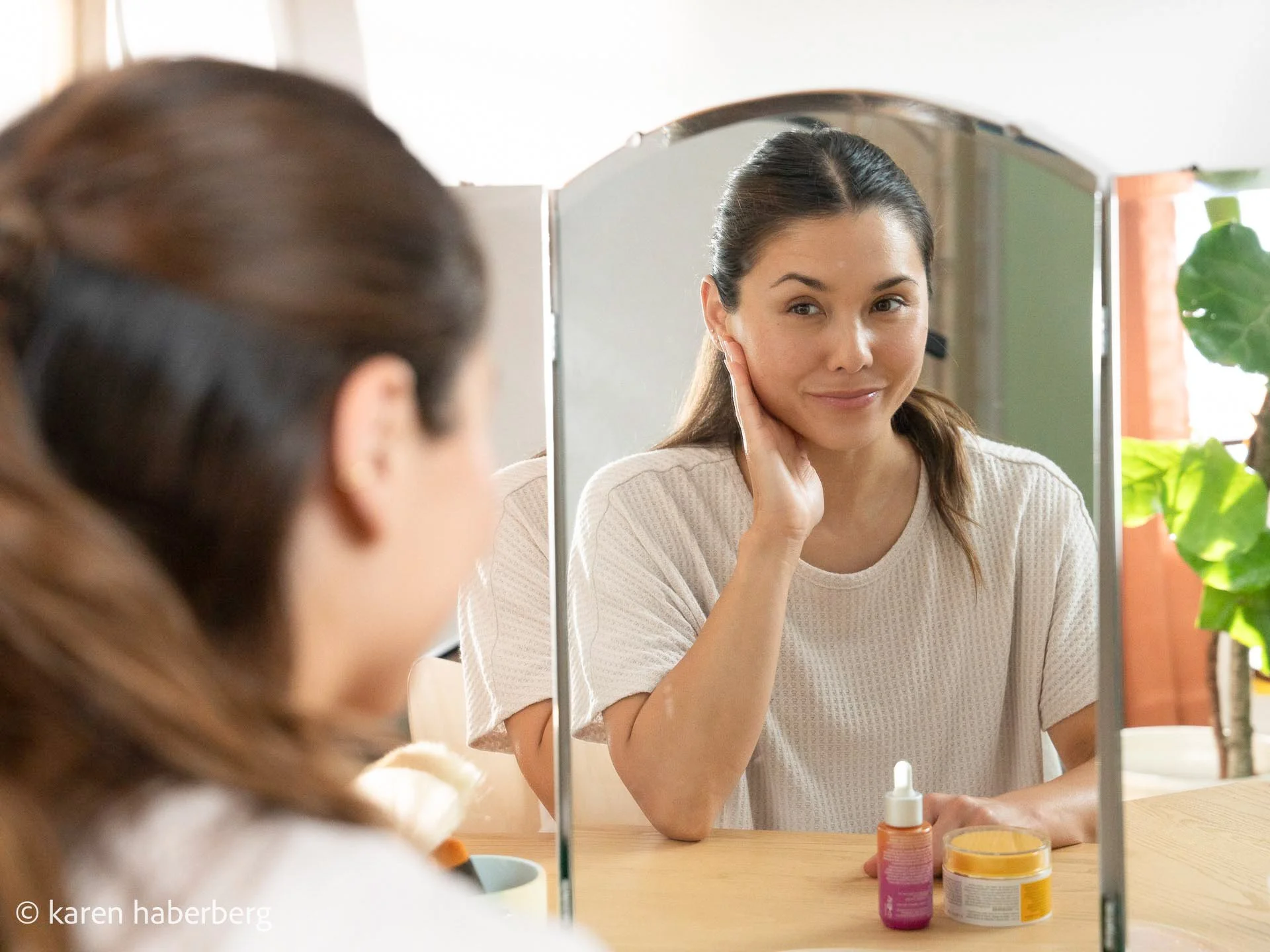 Woman looking at her reflection in a mirror, touching her neck, with skincare products on the table.