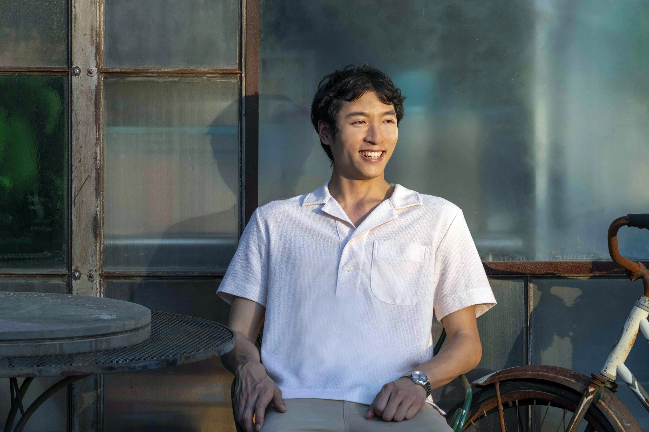 A young man with black hair and fair skin, wearing a white polo shirt, smiling while sitting outdoors next to a rusty bicycle and a round metal table, with an old industrial building in the background.