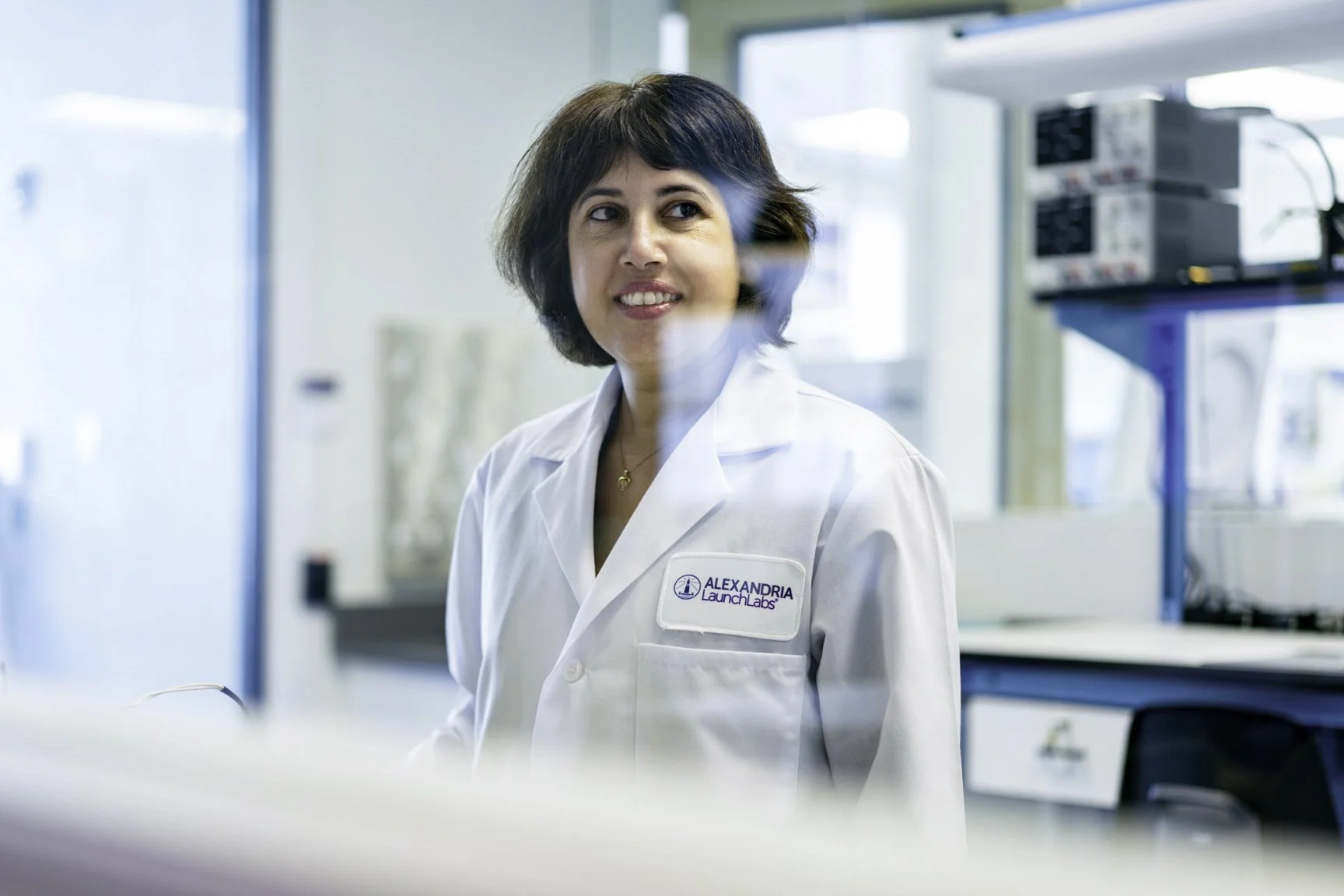 A woman wearing a white lab coat with the name tag 'Alexandria Launch Labs' inside a laboratory with scientific equipment in the background.