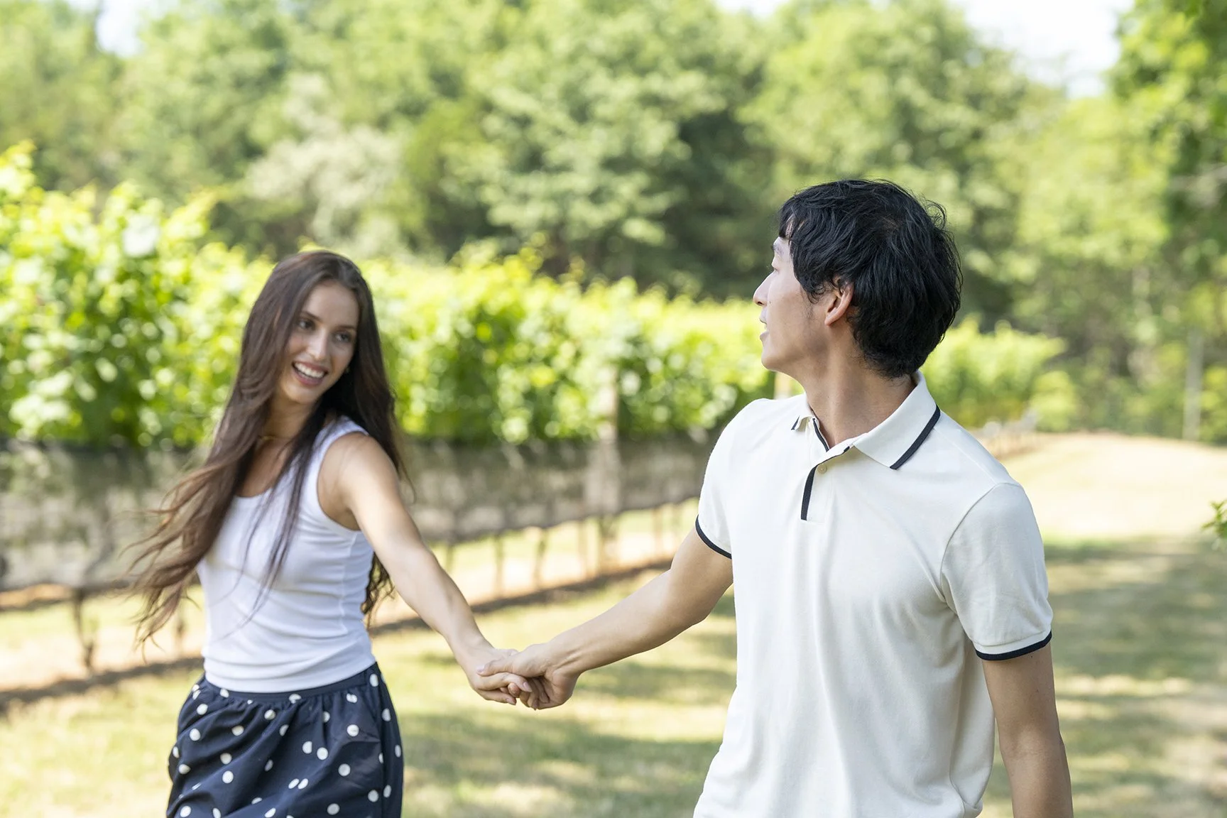 A man and a woman are holding hands and smiling at each other outdoors in a green park with trees in the background.