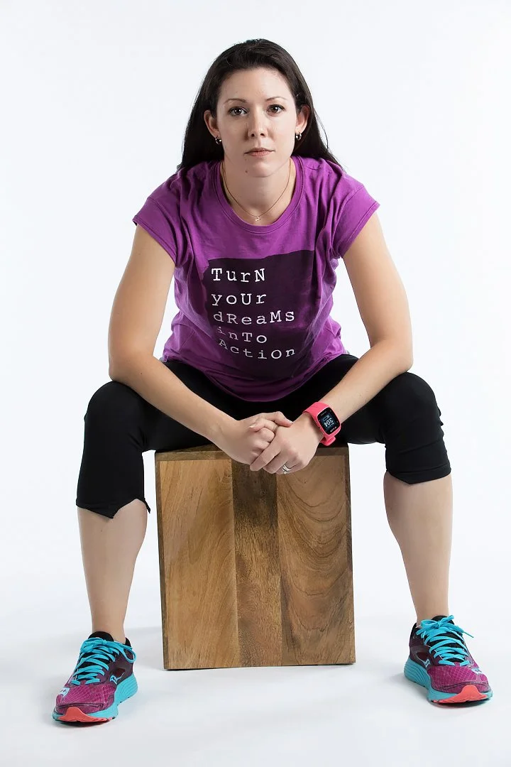 A woman with dark hair sitting on a wooden box, wearing a purple t-shirt with motivational text, black capris, colorful running shoes, and a sporty watch, against a plain white background.
