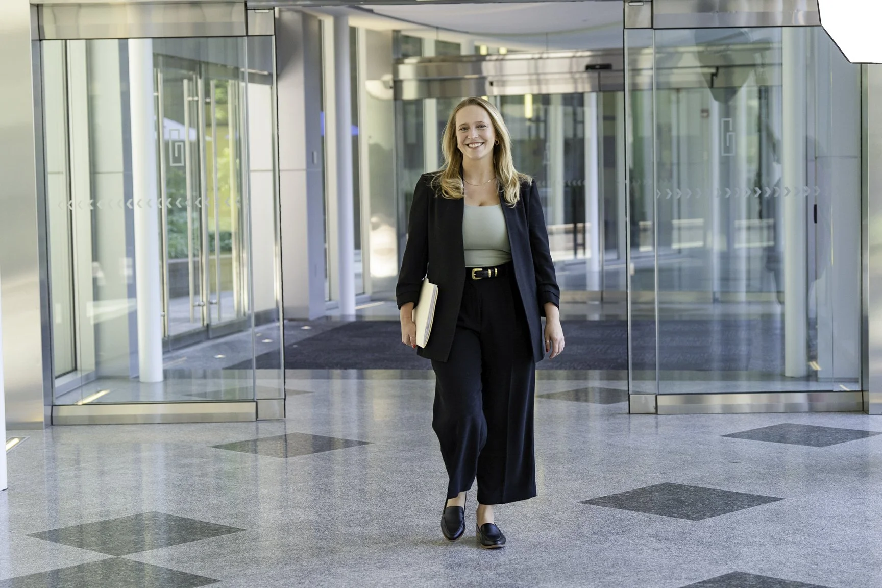 A woman in black pants, blazer, and loafers walking in the lobby of a modern office building with glass doors and windows while holding a folder.
