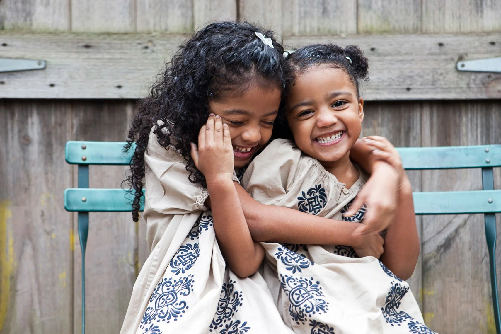 Two young girls smiling and hugging each other outdoors, sitting on a light blue metal bench with a wooden fence in the background.