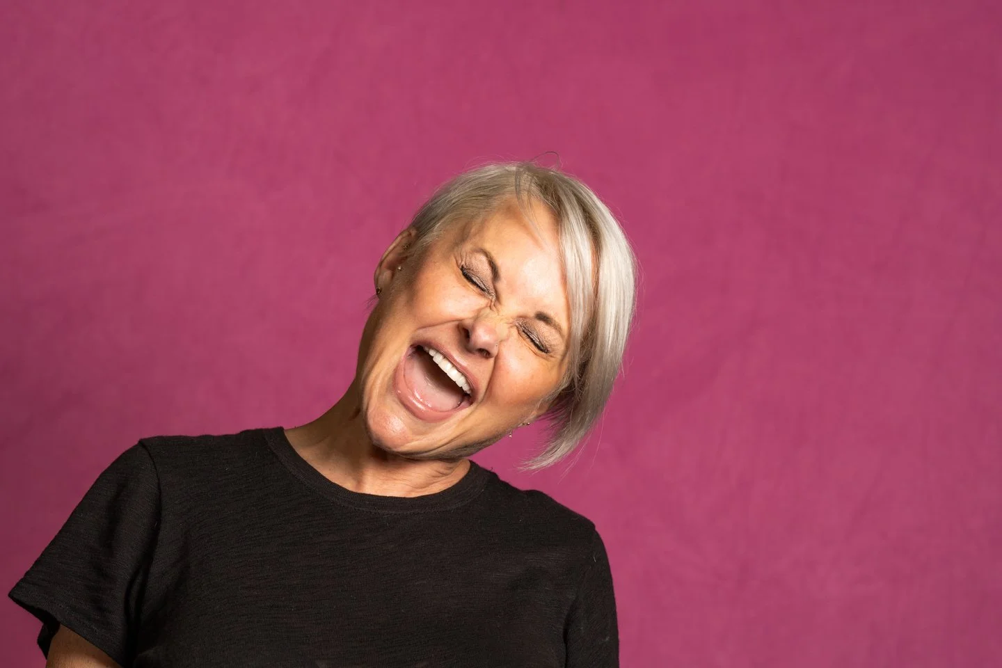 A woman with short gray hair singing with her eyes closed against a pink background.