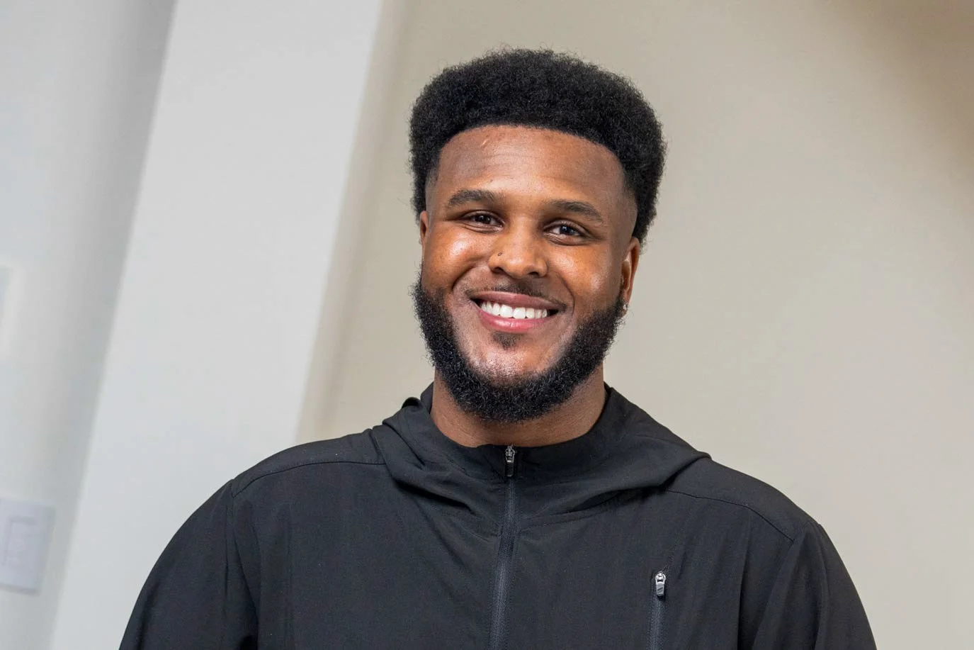 A smiling man with a beard and short curly hair, wearing a black zip-up jacket, standing indoors against a neutral background.