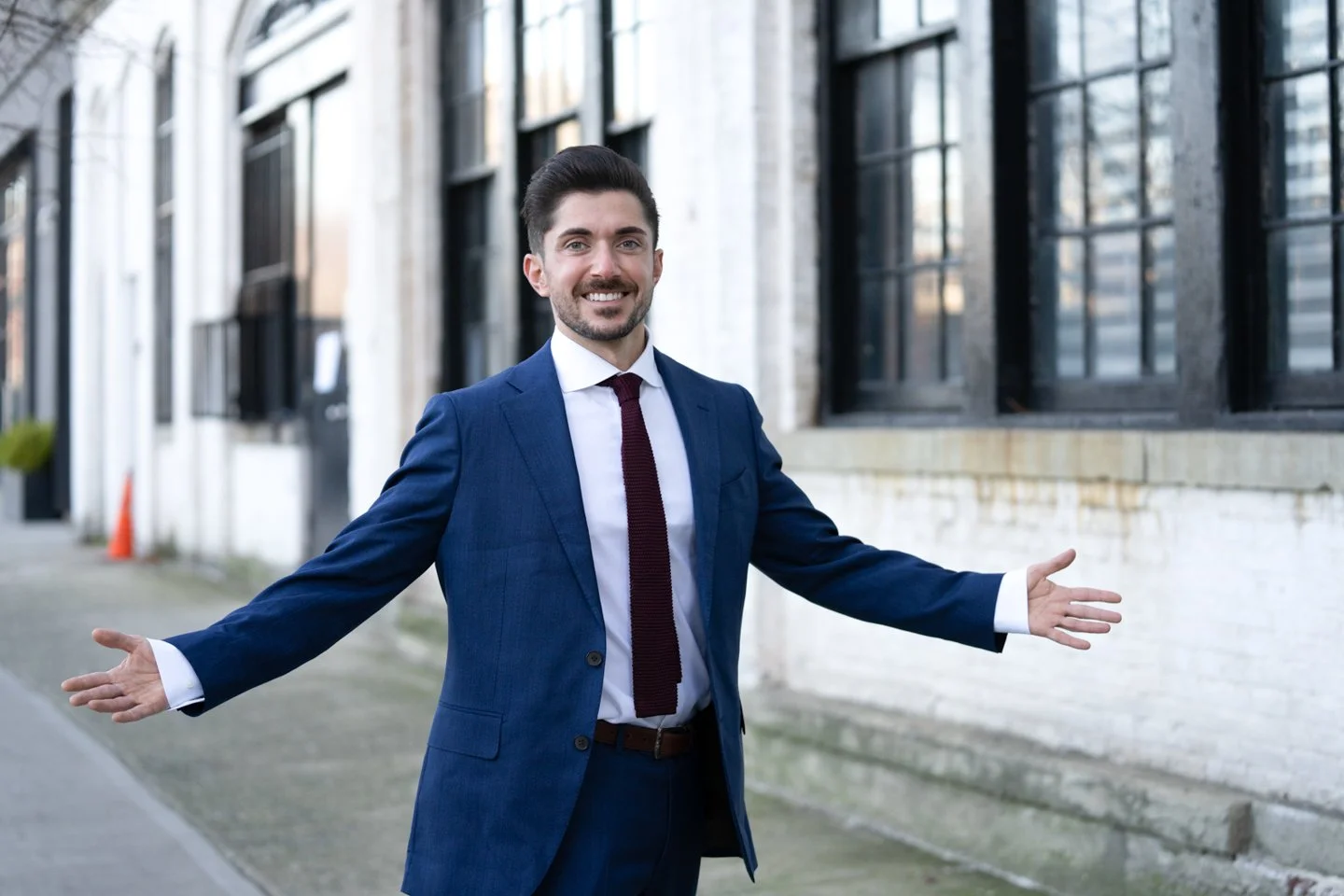 Man in a blue suit with a white shirt and dark red tie standing outdoors with open arms, smiling, next to a building with large windows.