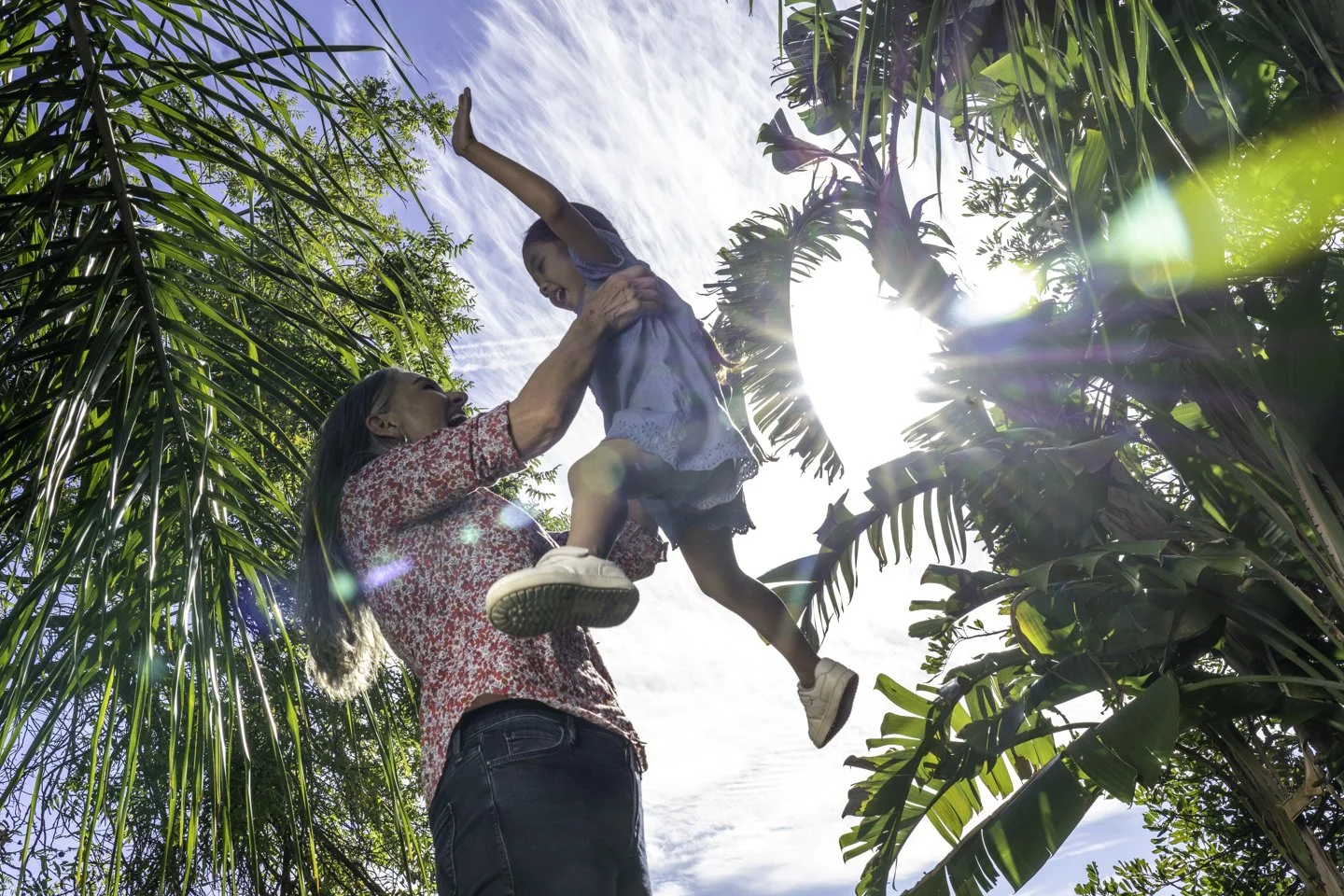 A woman lifts a young girl into the air outdoors among tall green trees and plants on a sunny day.