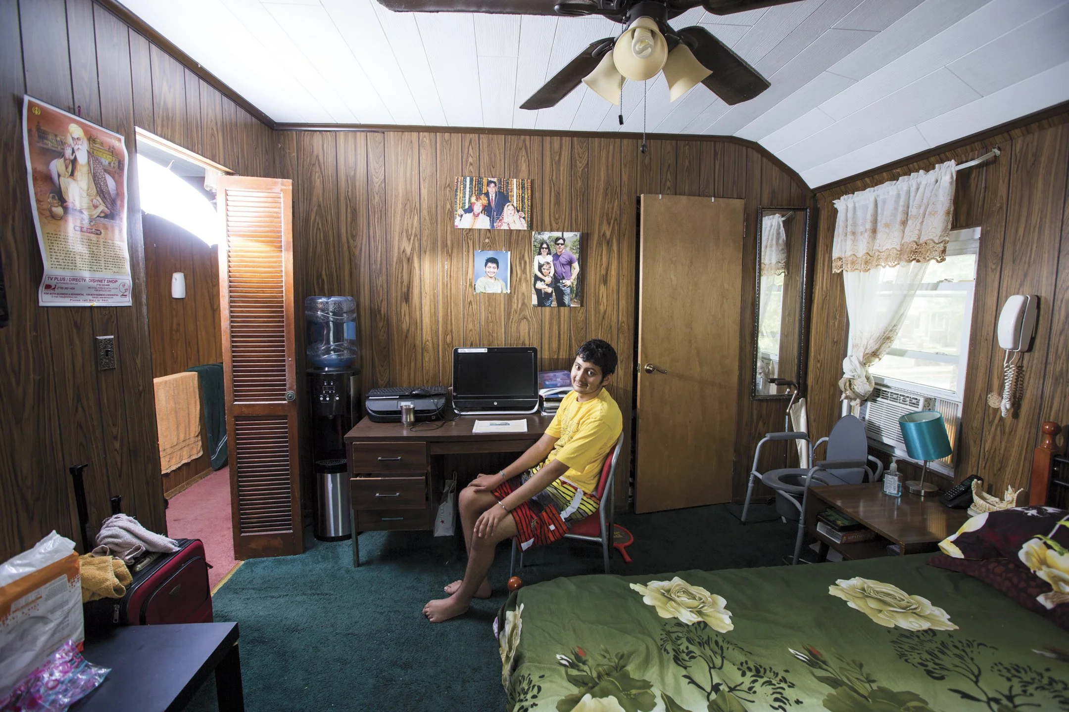 A young boy sitting in a small, wood-paneled bedroom with a green floral bedspread, a desk with a computer, and a water cooler. The room has a ceiling fan, curtains, and pictures on the wall.