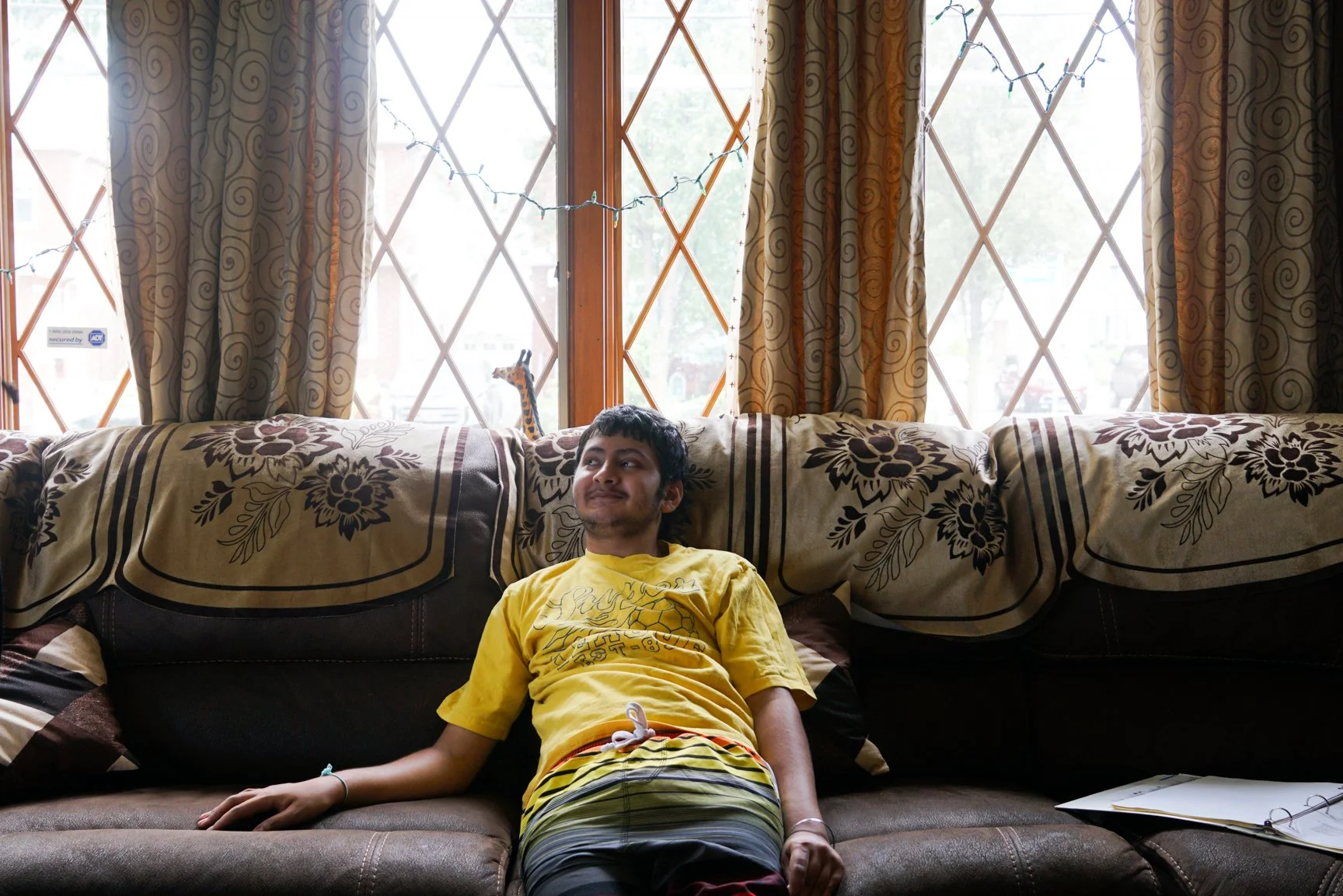Young man lying on a sofa in front of a large window with diamond-shaped panes, golden curtains, and string lights, with a small giraffe figurine visible behind him.