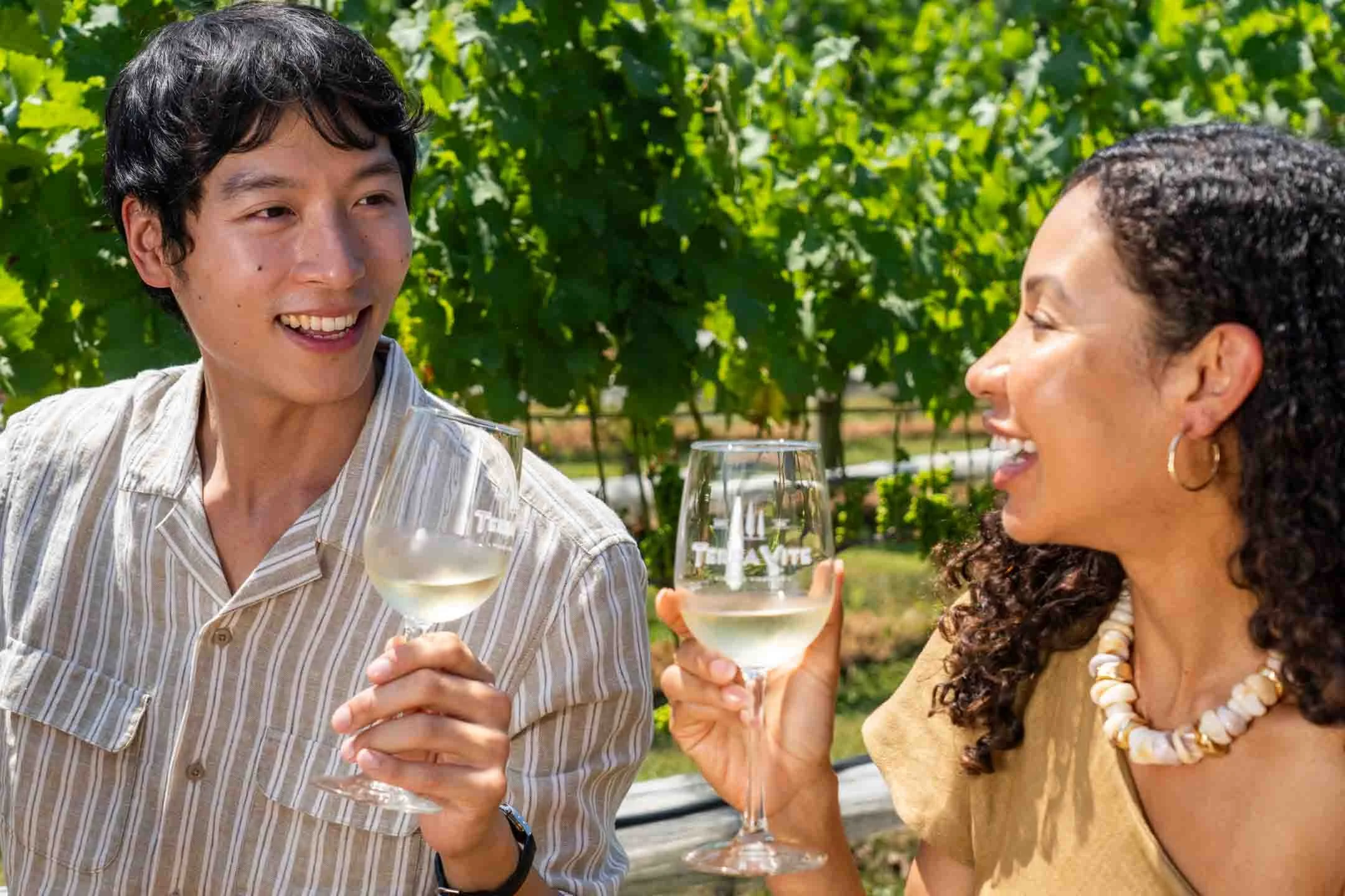 A young man and woman smiling and toasting with glasses of white wine outdoors on a sunny day, with green foliage in the background.