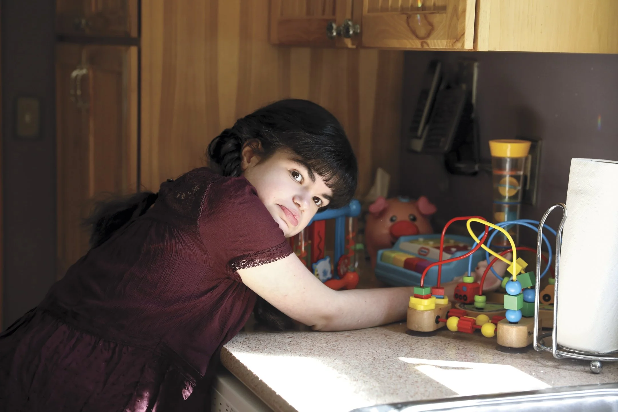 A young girl with black hair and a maroon dress playing with a bead maze toy on a kitchen counter, smiling at the camera.