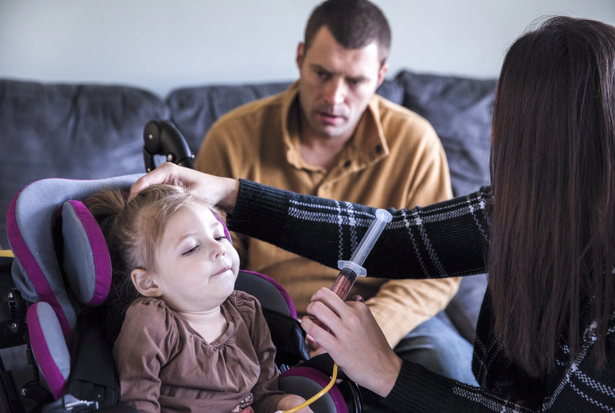 A young girl sitting in a car seat being administered oral medicine by a woman while a man watches, in a living room.