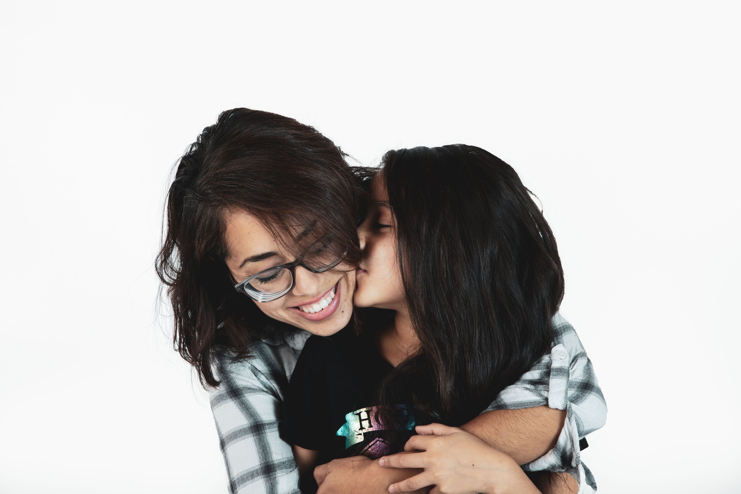 A woman with glasses hugging and smiling at a young girl, who is giving her a kiss on the cheek, against a plain white background.