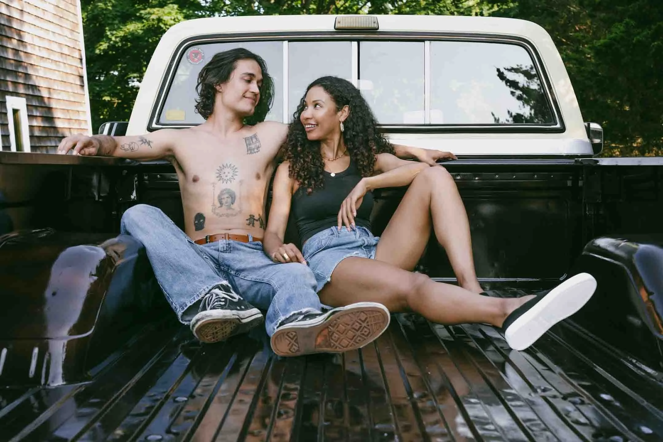 A man and woman sitting together in the bed of a pickup truck outdoors, looking at each other and smiling, with trees and a house visible in the background.