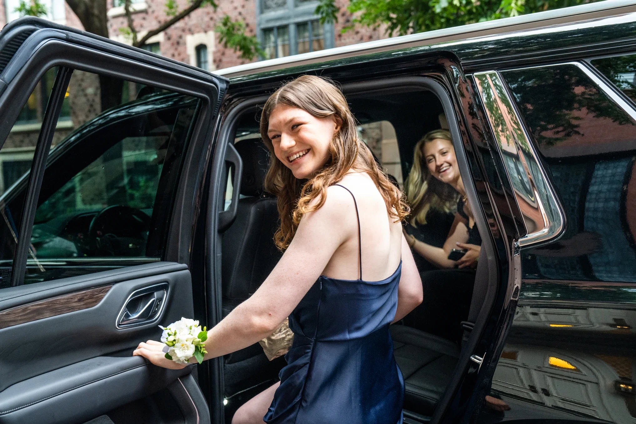 Young woman in a navy blue dress with a white corsage, smiling as she steps out of a black car, with another woman in the backseat also smiling.
