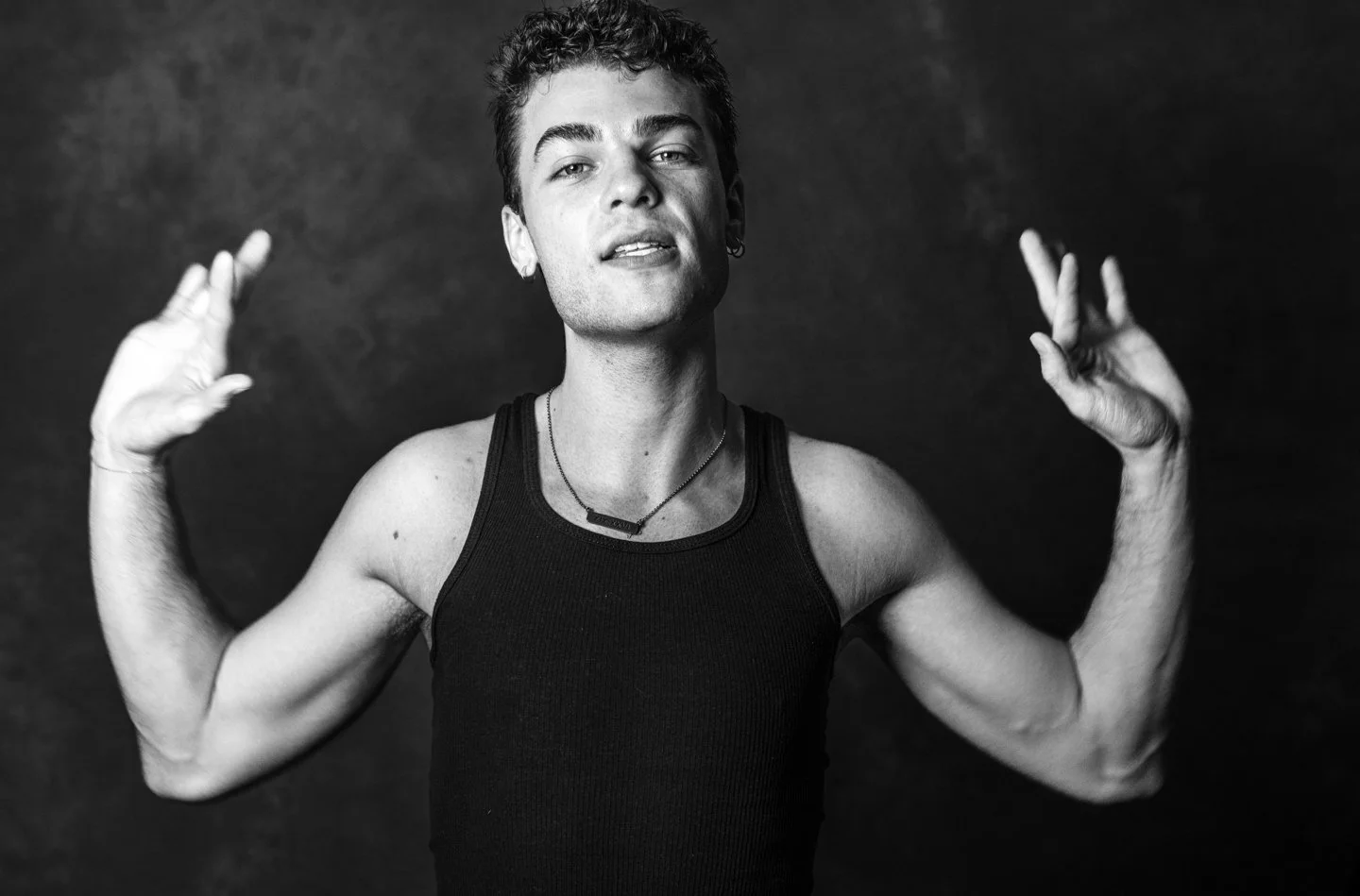Black and white portrait of a young man with short curly hair, wearing a black tank top and a necklace, with both hands raised near his head, against a dark background.