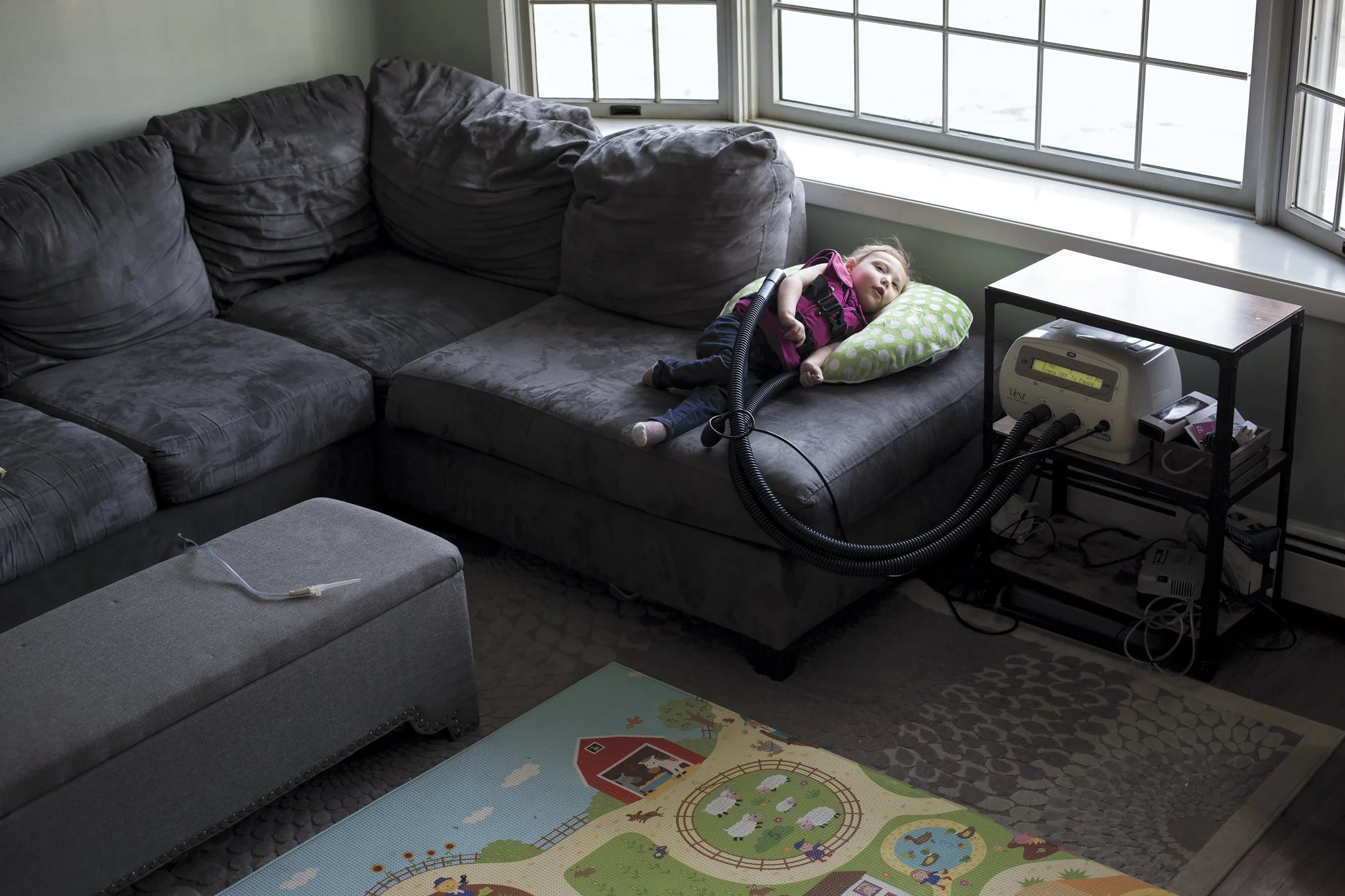 A young child lying on a gray sectional sofa with a green polka-dot pillow, connected to a medical ventilator on a nearby black metal table in a living room with a large window.
