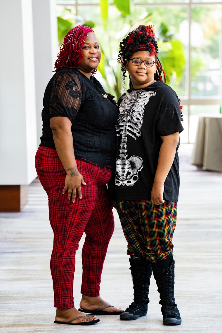 Two women standing inside in front of a window with green plants visible outside. One woman has red dreadlocks and is wearing a black lace top and red plaid pants. The other woman has curly hair with red highlights, glasses, and is wearing a black t-