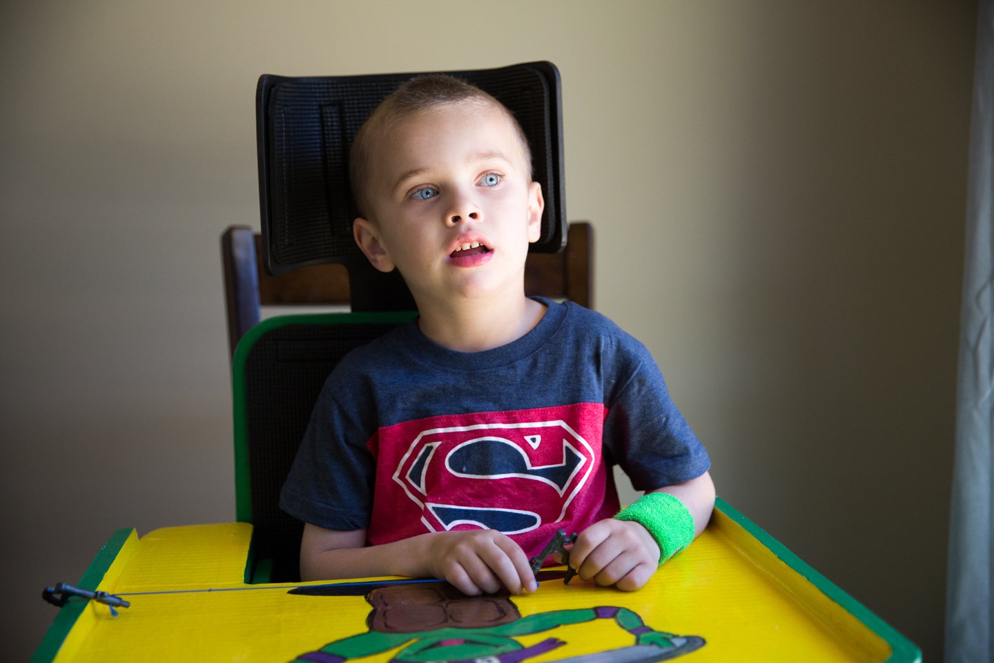 A young boy with blue eyes and short light brown hair sitting at a yellow table with a Superman shirt, holding a fishing rod, wearing a green wristband, in a room with a beige wall and a window with a white curtain.