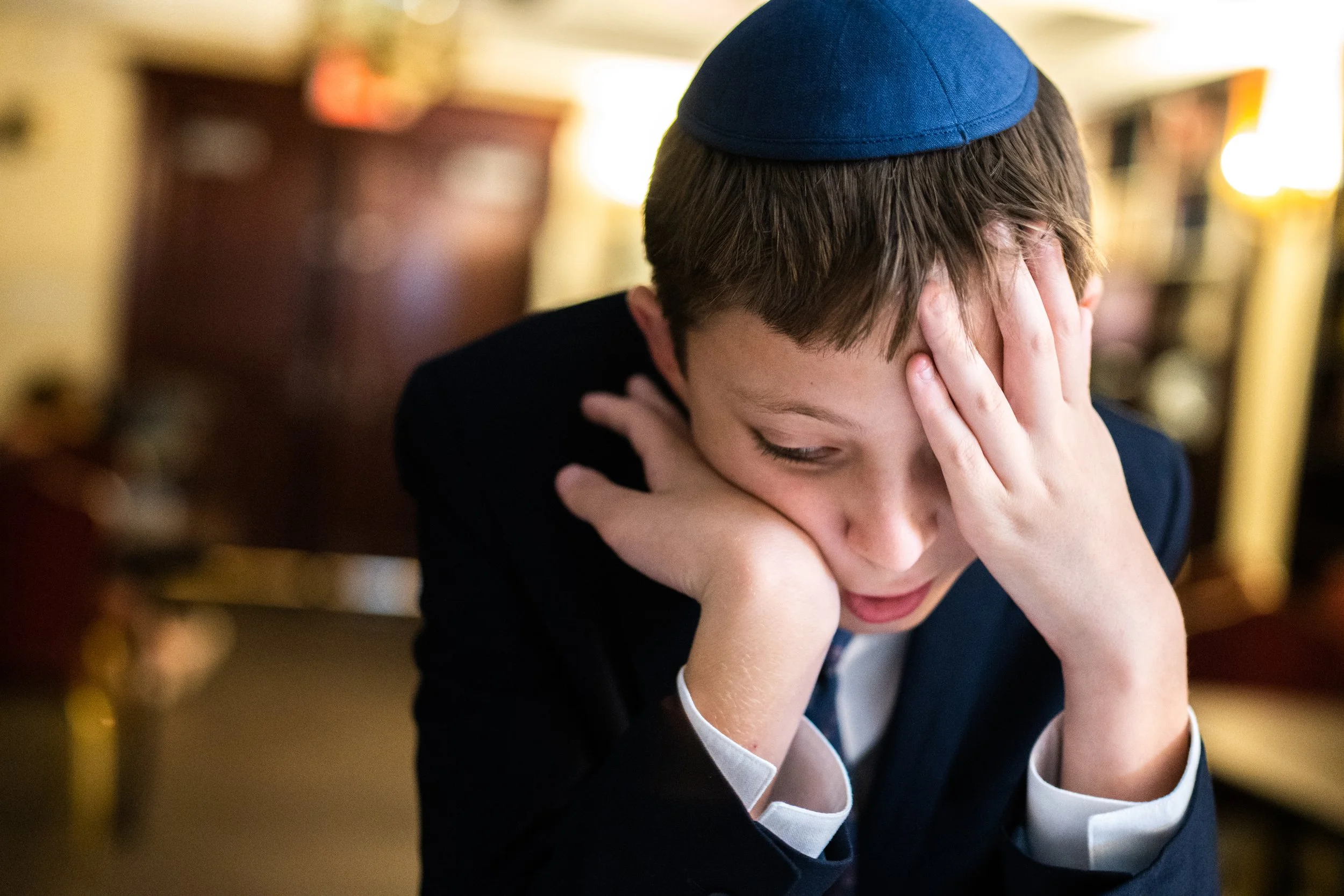 A young boy wearing a blue yarmulke, dressed in formal attire, is holding his head with both hands and appears distressed or worried.