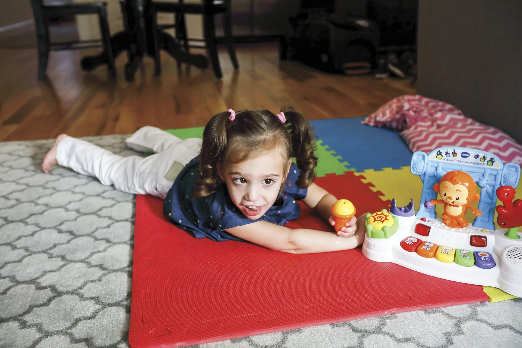 Young girl lying on her stomach on a colorful foam mat playing with a electronic toy set featuring a monkey and colorful buttons.