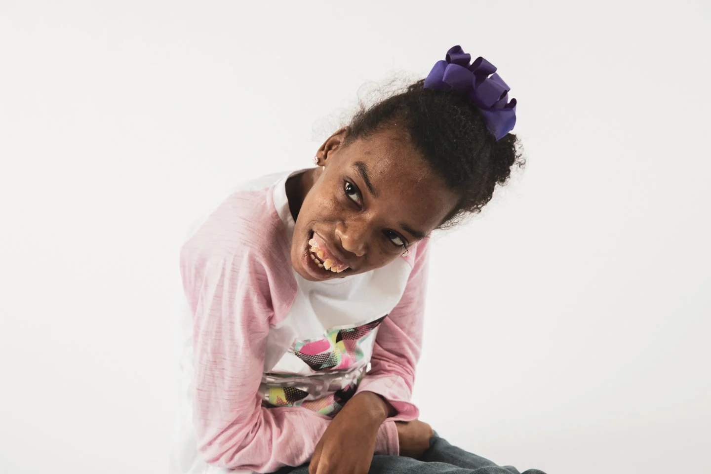 Young girl with a purple bow in her hair, wearing a pink and white long-sleeve shirt, smiling and leaning forward with her mouth open against a plain white background.