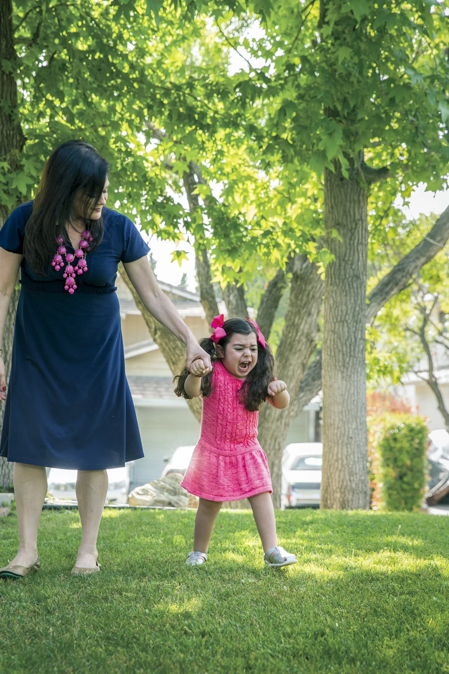 A woman in a navy blue dress and pink necklace is holding hands with a young girl in a pink dress, who appears upset or crying, on a grassy lawn under trees in a suburban neighborhood.