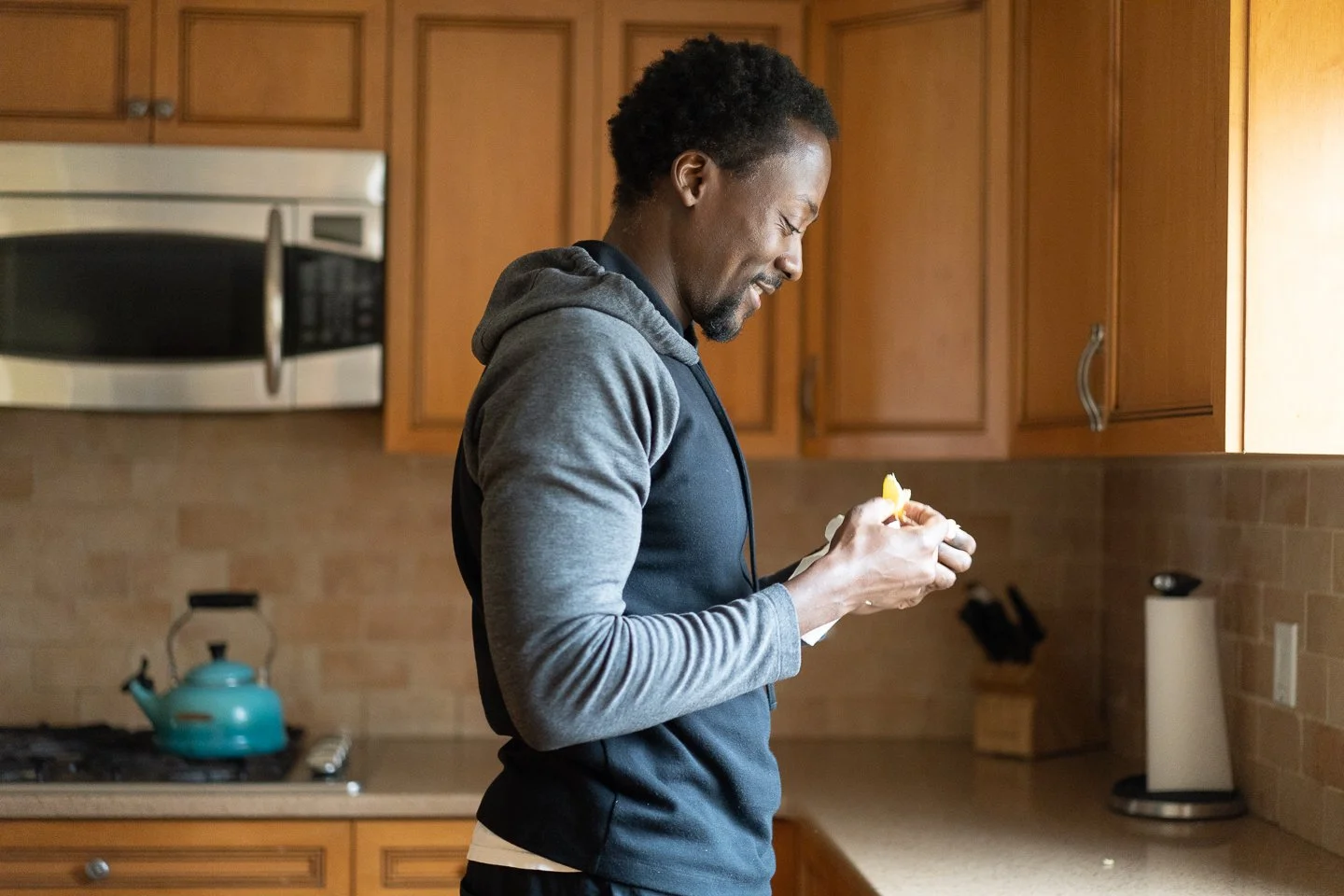 A man wearing a black and gray hoodie in a kitchen, smiling and looking at something in his hands.