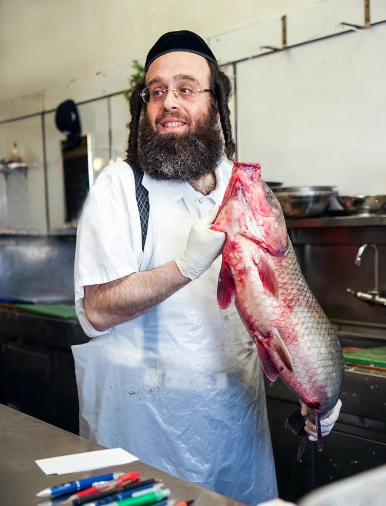 A man with glasses, a beard, and curly hair wearing a white apron, black skullcap, and gloves, holds a large fish by the head in a kitchen or fish market setting.