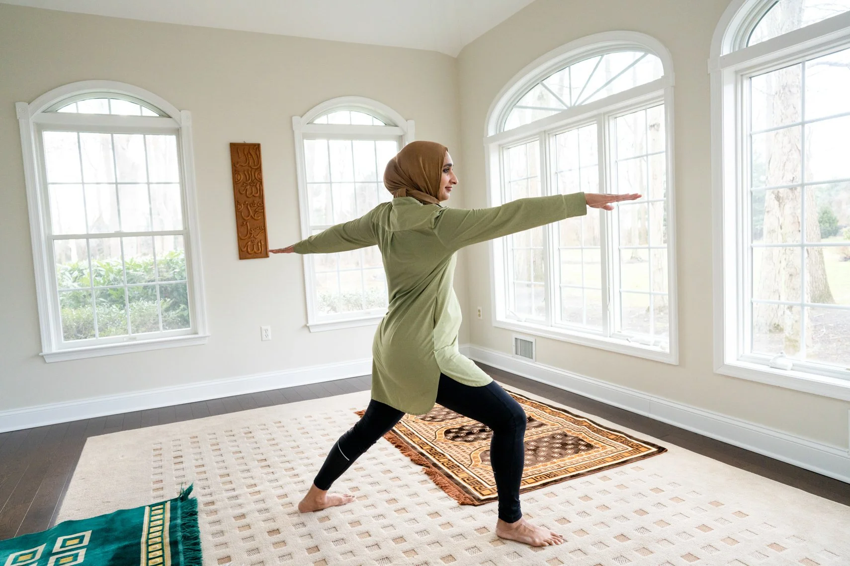A woman in modest clothing, including a hijab, practicing yoga in a bright room with large arched windows and patterned rugs.