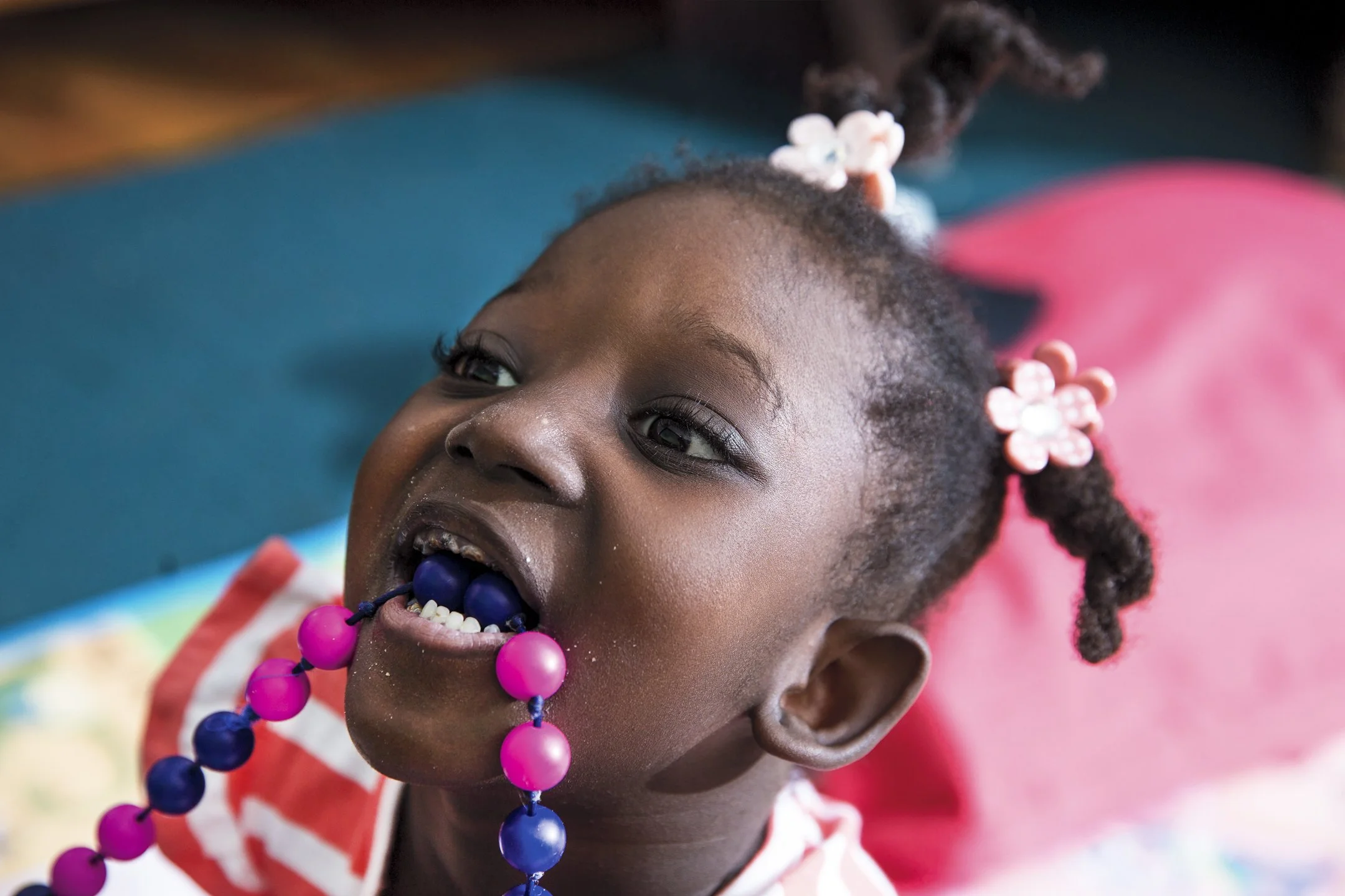 A young girl with braided hair decorated with pink flower clips, lying on her back, playing with colorful beads in her mouth, and wearing a red and white striped shirt.