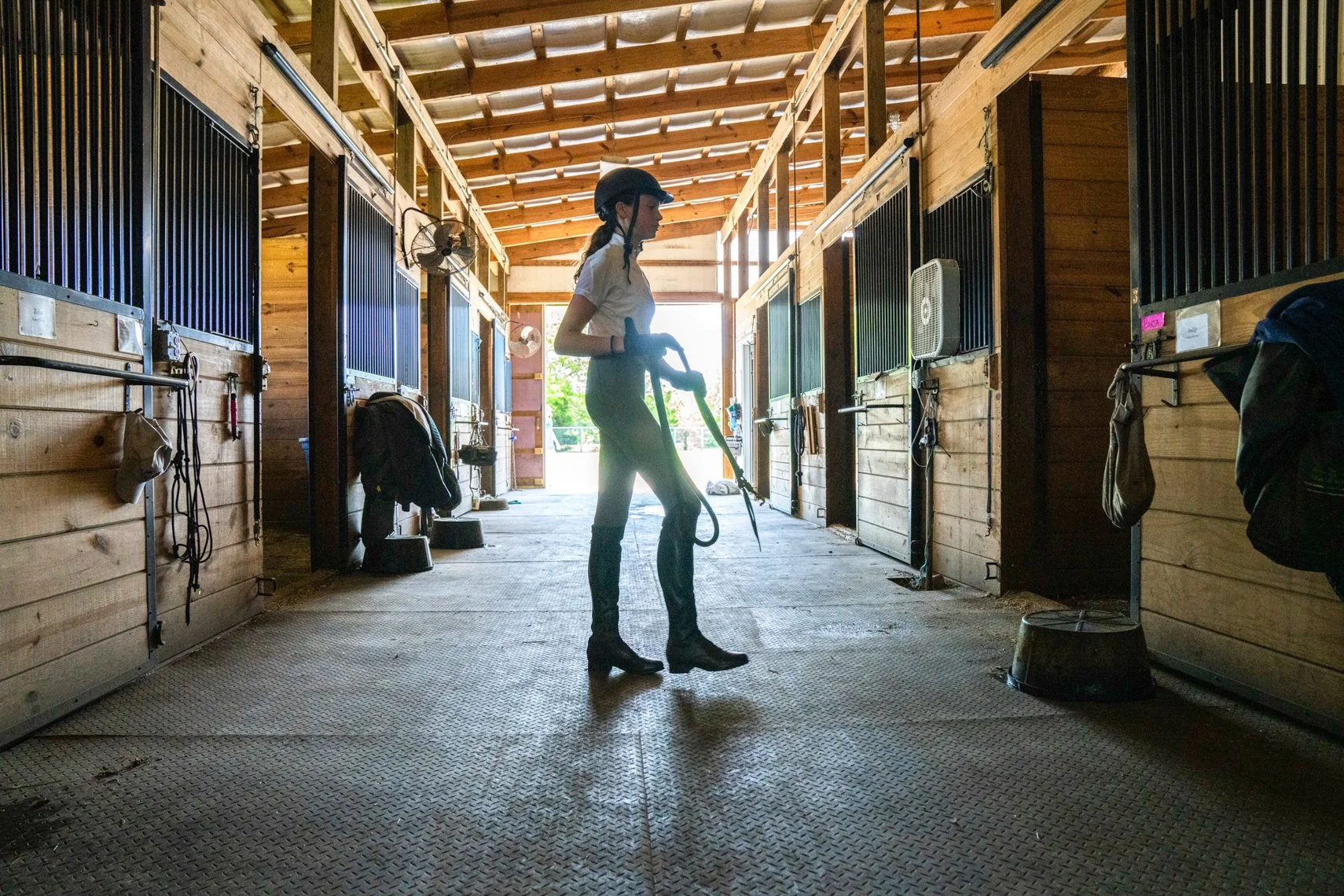 A woman cleaning horse stalls in a wooden barn with a vacuum cleaner.