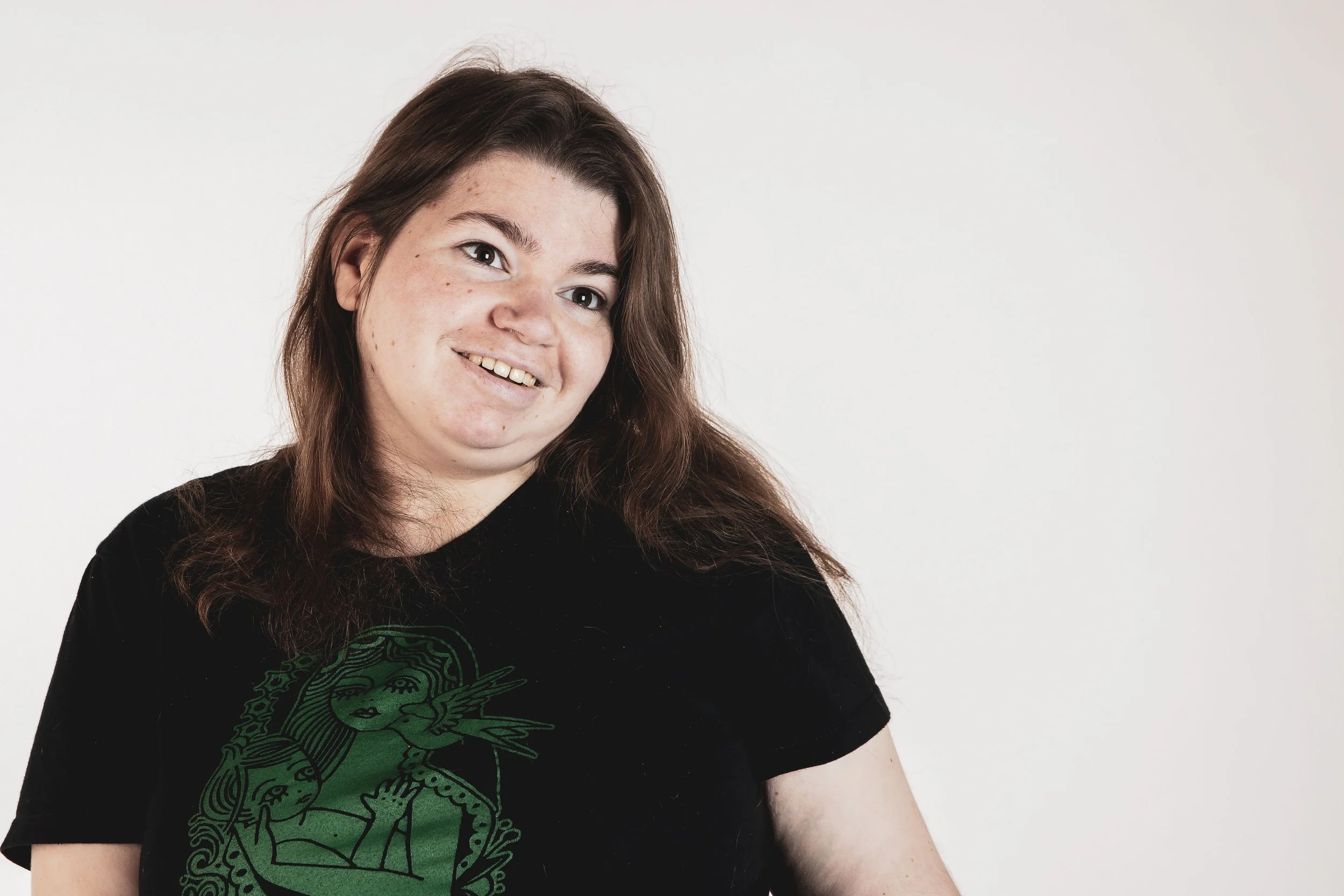 A young woman with long brown hair smiling, wearing a black t-shirt with a green graphic design of a woman with wings, against a plain white background.