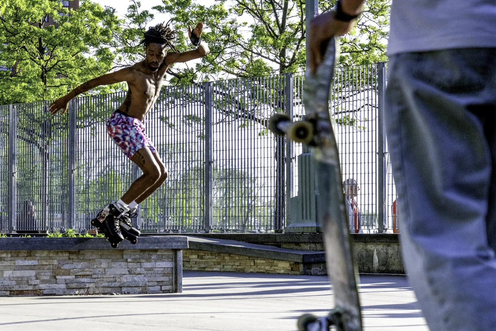 A young man is rollerblading on a low ledge at a skatepark while another person holds a skateboard nearby. There is green foliage and a metal fence in the background.