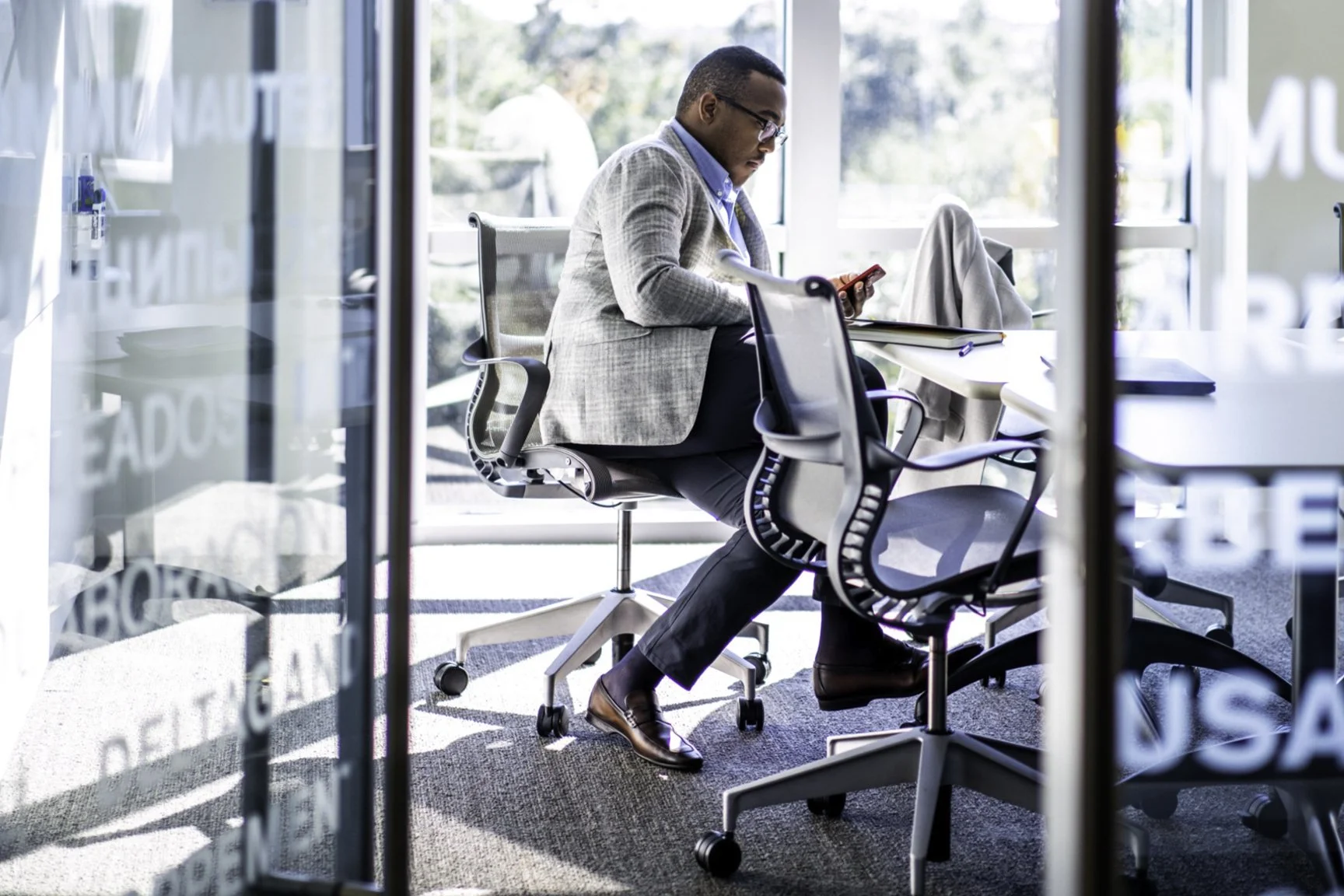 Man in a gray blazer and glasses sitting at a conference table, looking at his phone in a bright, modern office with large windows.