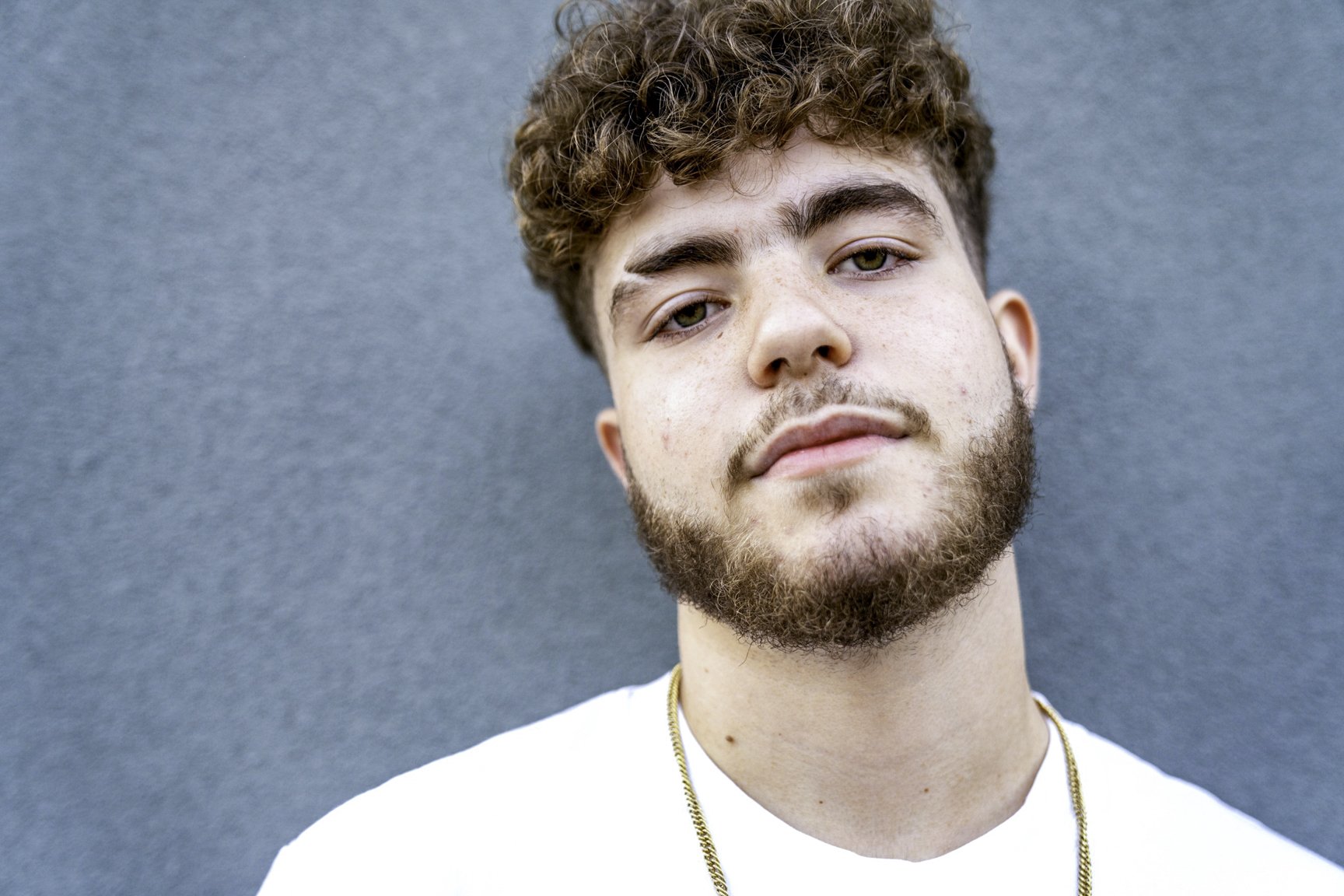 Close-up portrait of a young man with curly brown hair and a beard, wearing a white shirt and a gold chain, against a blue background.