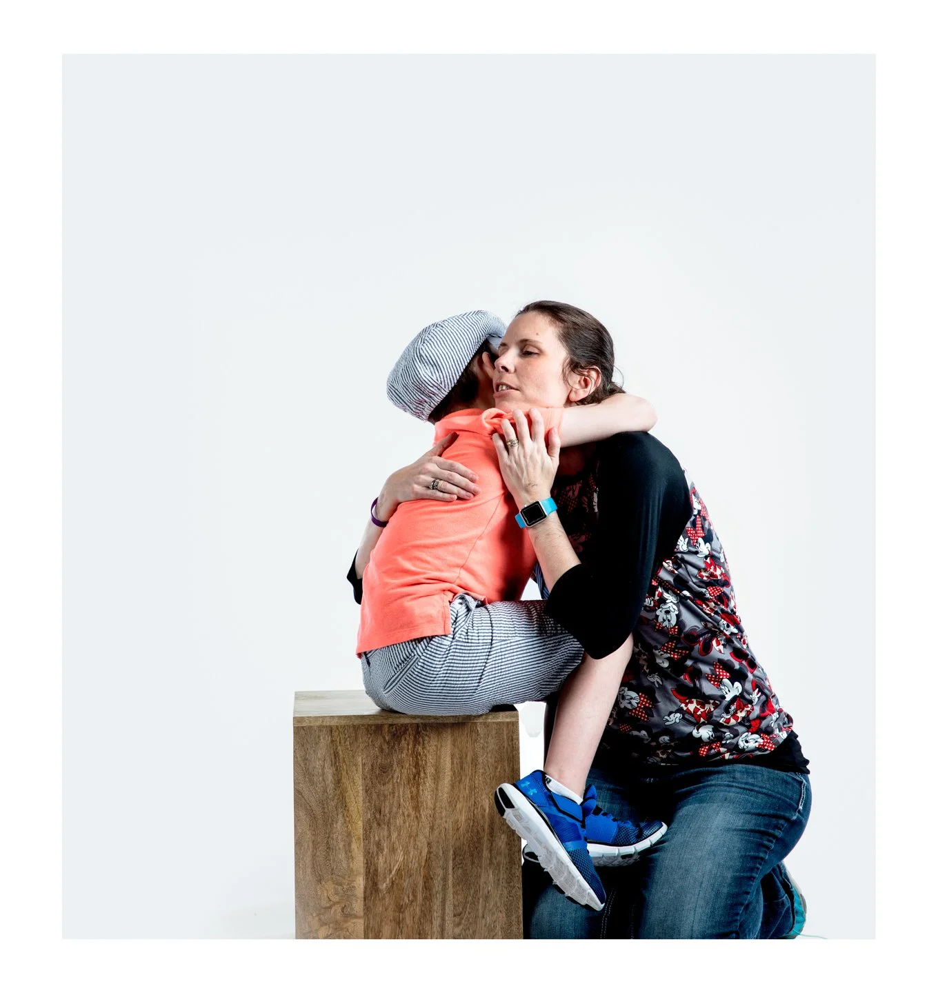 A woman with dark hair hugging a child who is sitting on a wooden block. The child is wearing a gray hat, pink shirt, gray striped pants, and blue shoes. The woman is wearing a black and patterned dress and has an Apple Watch on her wrist.